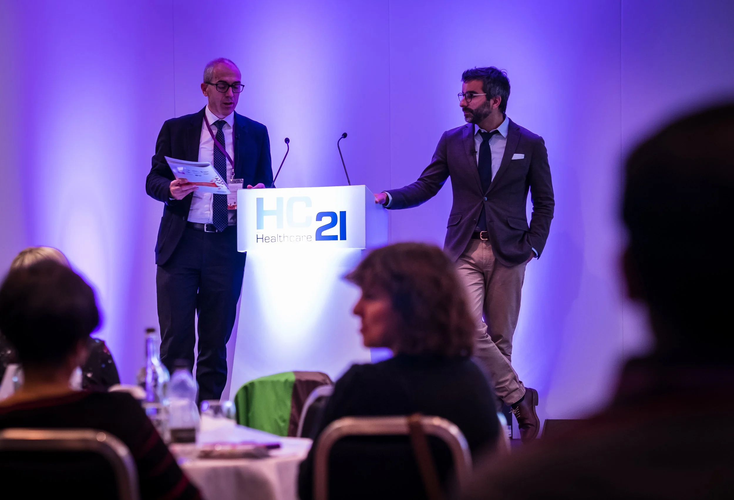 Two men in suits on a stage at a healthcare conference, one holding papers, with an audience in front and a sign that says 'HC21 Healthcare'.