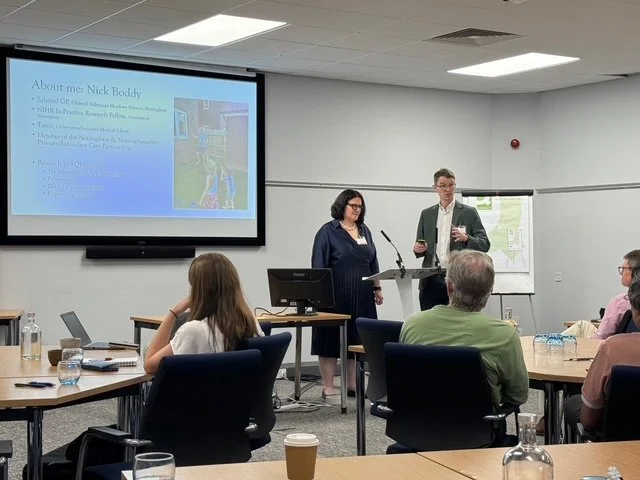 A professional presentation in a conference room with a woman and a man speaking at the front. The slide on the screen introduces Nick Boddy and contains a photo and bio. Attendees are seated facing the speakers, some taking notes or using laptops.