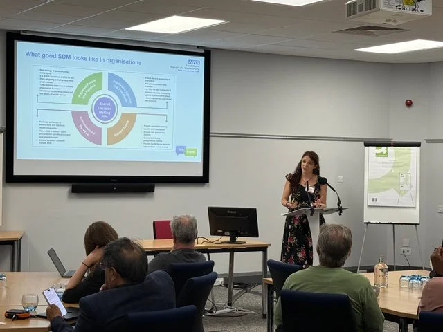 A woman giving a presentation in a conference room with a large screen displaying a slide titled 'What good SDM looks like in organisations.' The slide features a circular diagram with text, and there are attendees seated listening, some taking notes