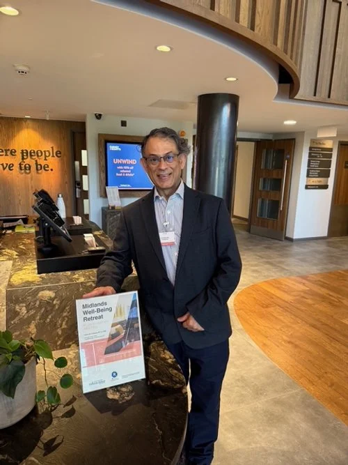 A man in a suit standing at a reception desk, smiling, with a sign for the Midlands Well-Being Retreat on the counter.