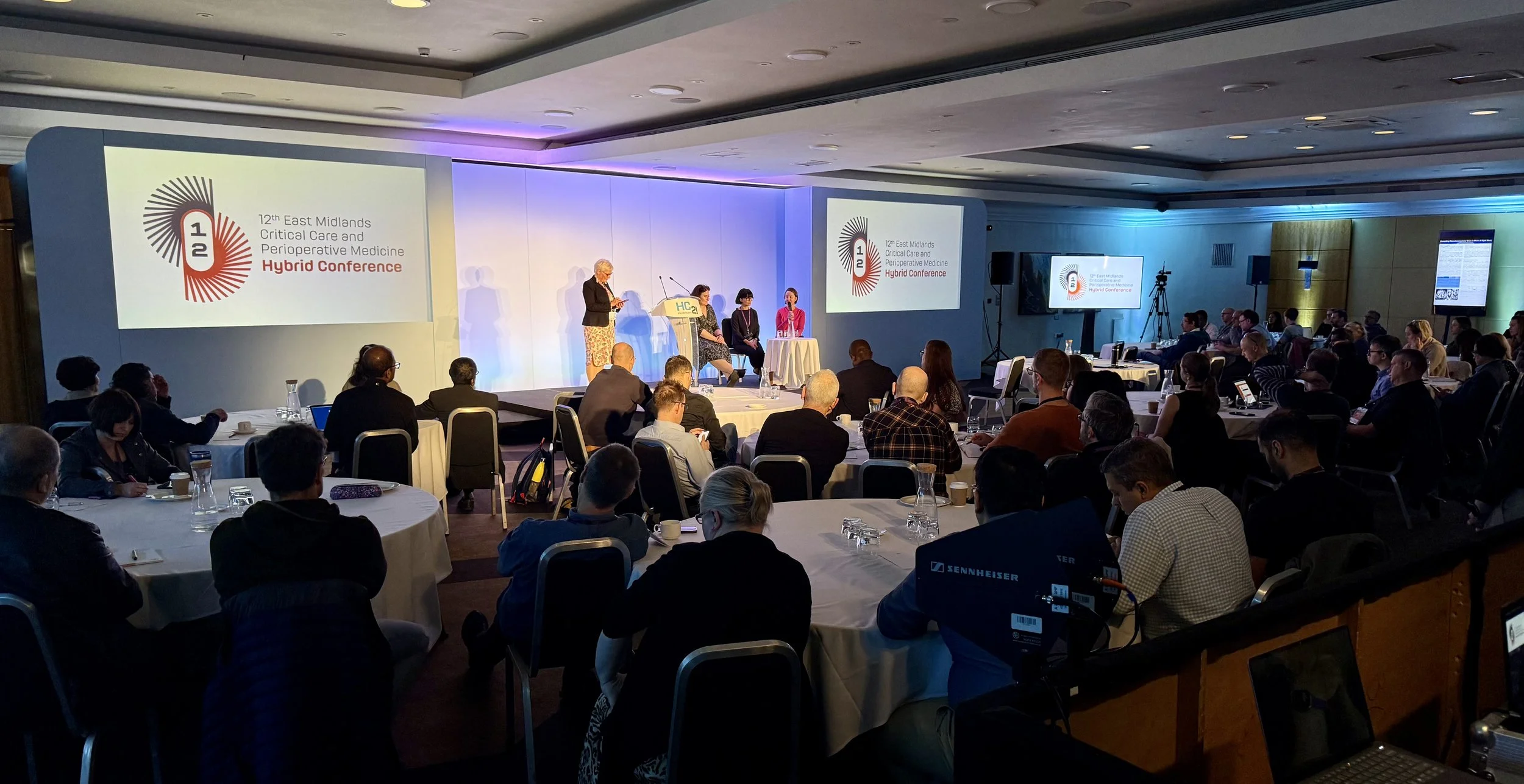 Conference room with a stage and a large screen displaying event logo, audience seated at round tables, speaker at microphone on stage, panel members seated nearby, and multiple screens showing event information.