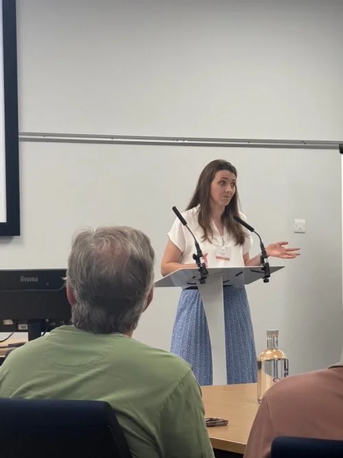 Woman giving a presentation at a podium with a microphone, in a conference room with an audience.