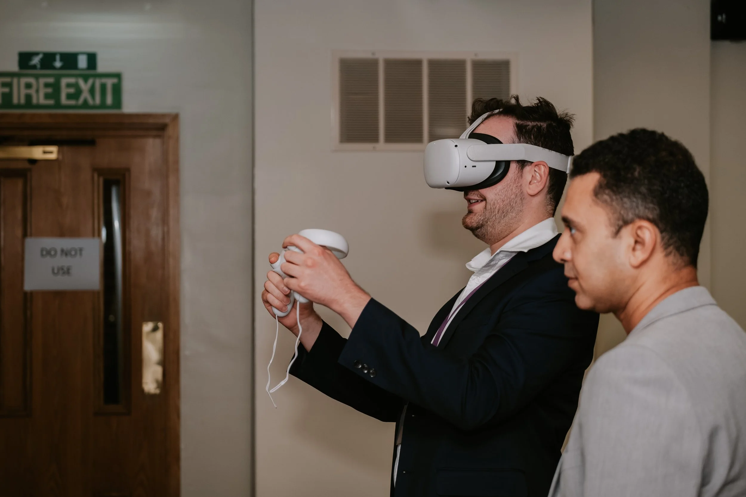 Two men are standing indoors, one man wearing a virtual reality headset and holding a handheld controller, while the other man watches. The man with the headset is smiling and engaging in a virtual experience.