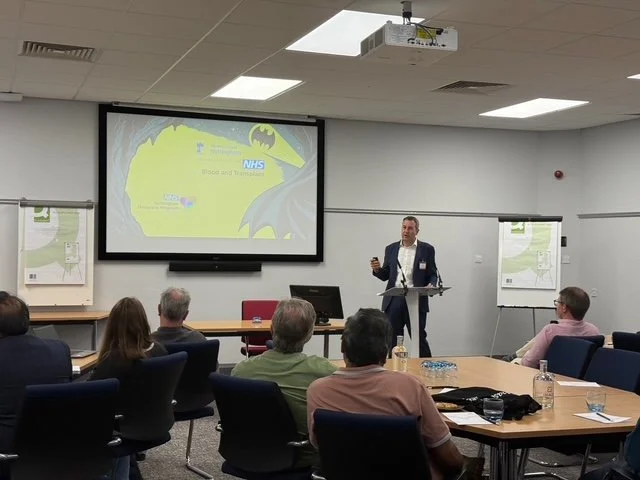 A man presenting at a conference or meeting room with a large screen displaying Batman-themed graphics and logos.