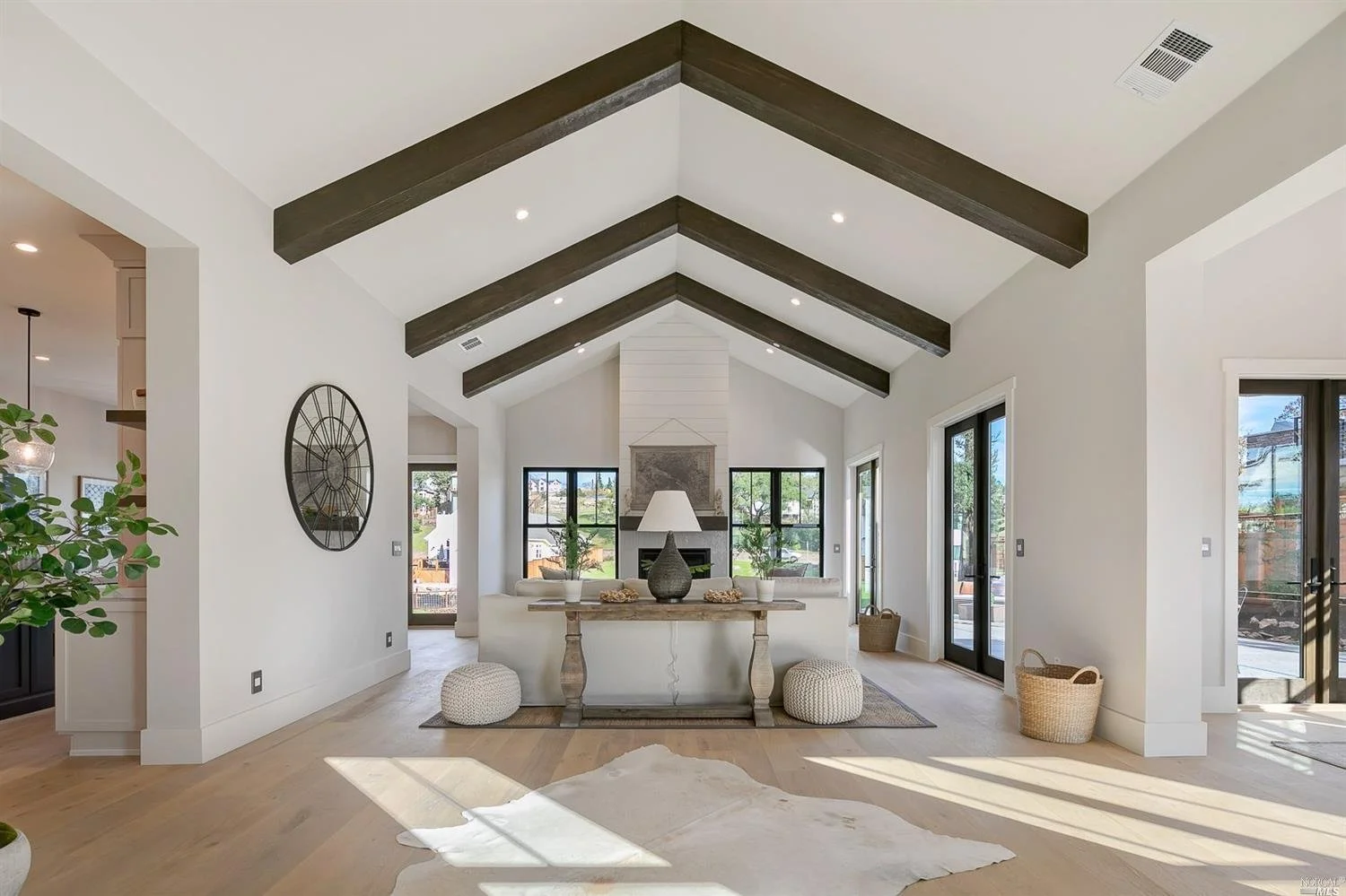 Bright living room with high vaulted ceiling featuring dark wooden beams, large windows, and sliding glass doors, with light-colored walls and hardwood flooring, and a fireplace with a white mantel.