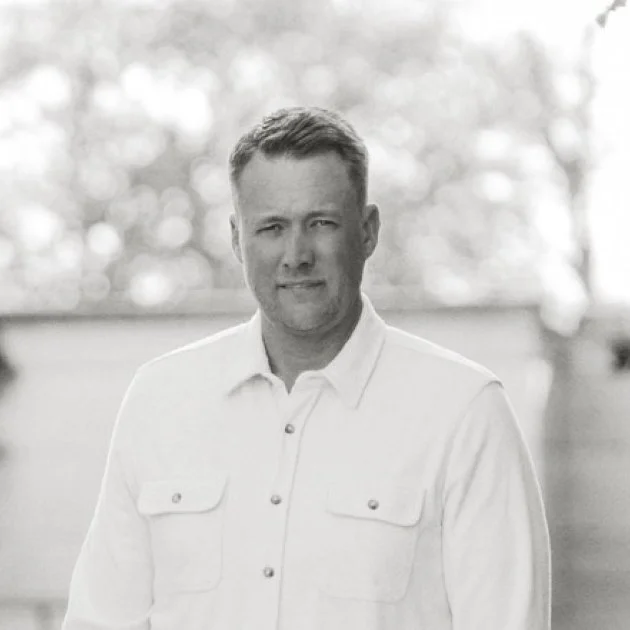 A black and white photo of a man wearing a light-colored button-up shirt, standing outdoors with blurred trees in the background.