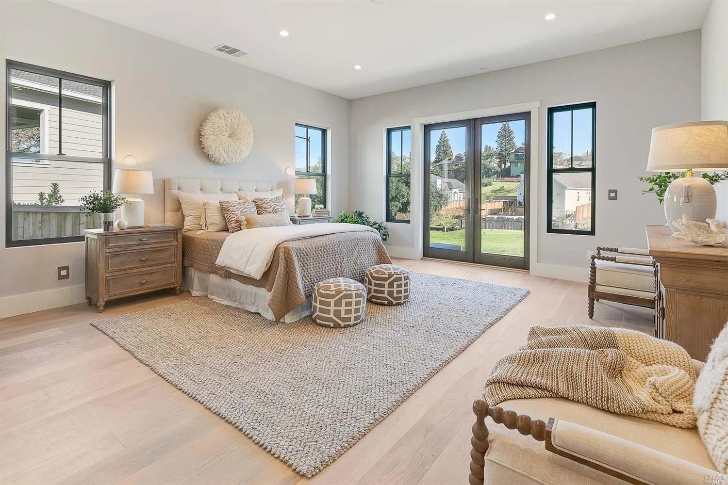 Bright bedroom with a large bed, beige and white bedding, two ottomans at the foot, nightstands with lamps on either side, a beige rug, a wooden bench, colorful pillows, and large windows and glass doors showing an outdoor view with houses and greenery.