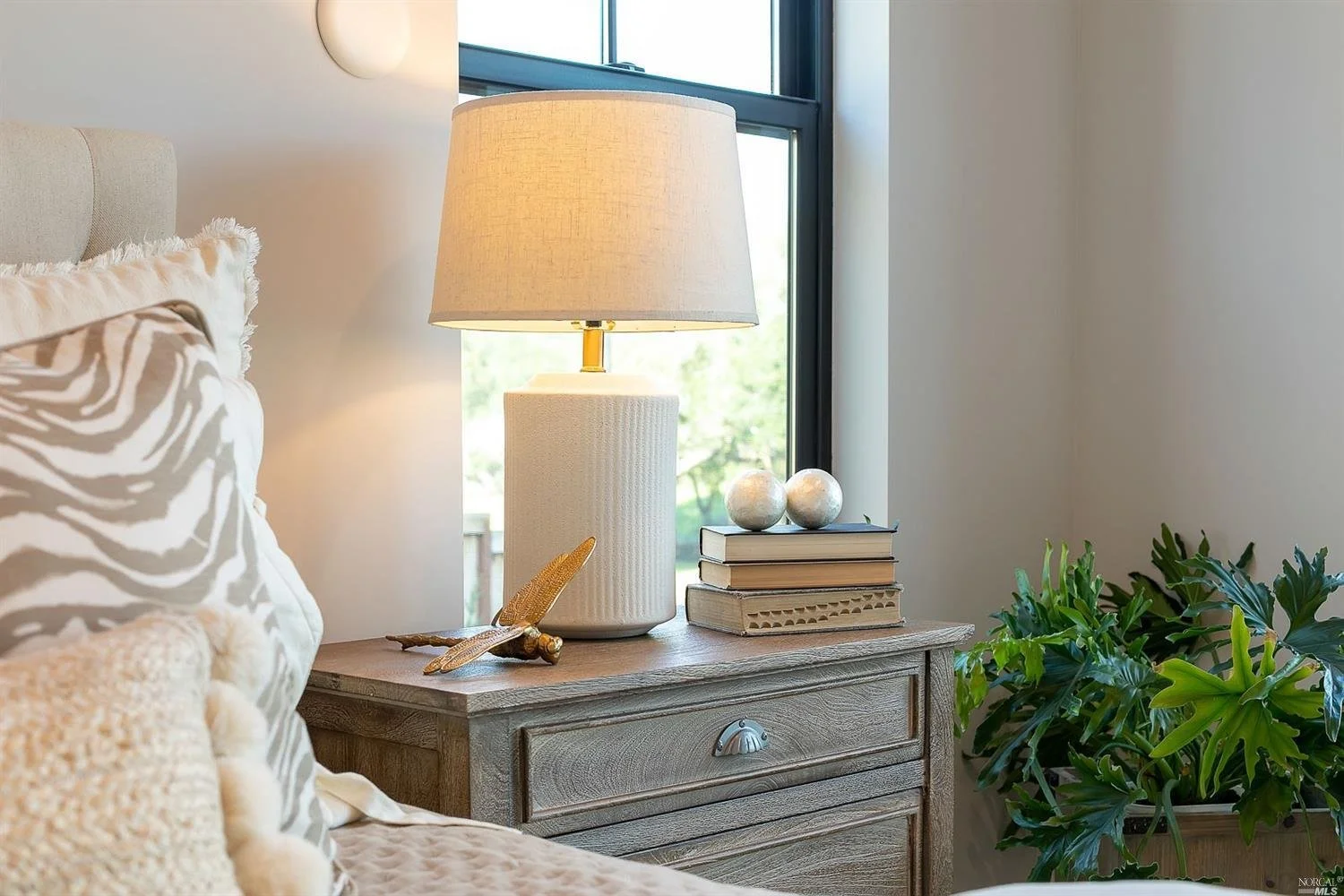 A cozy bedroom nightstand with a table lamp, decorative books, metallic spheres, a leaf-shaped ornament, and a plush pillow, near a window with a view of trees and a house outside.