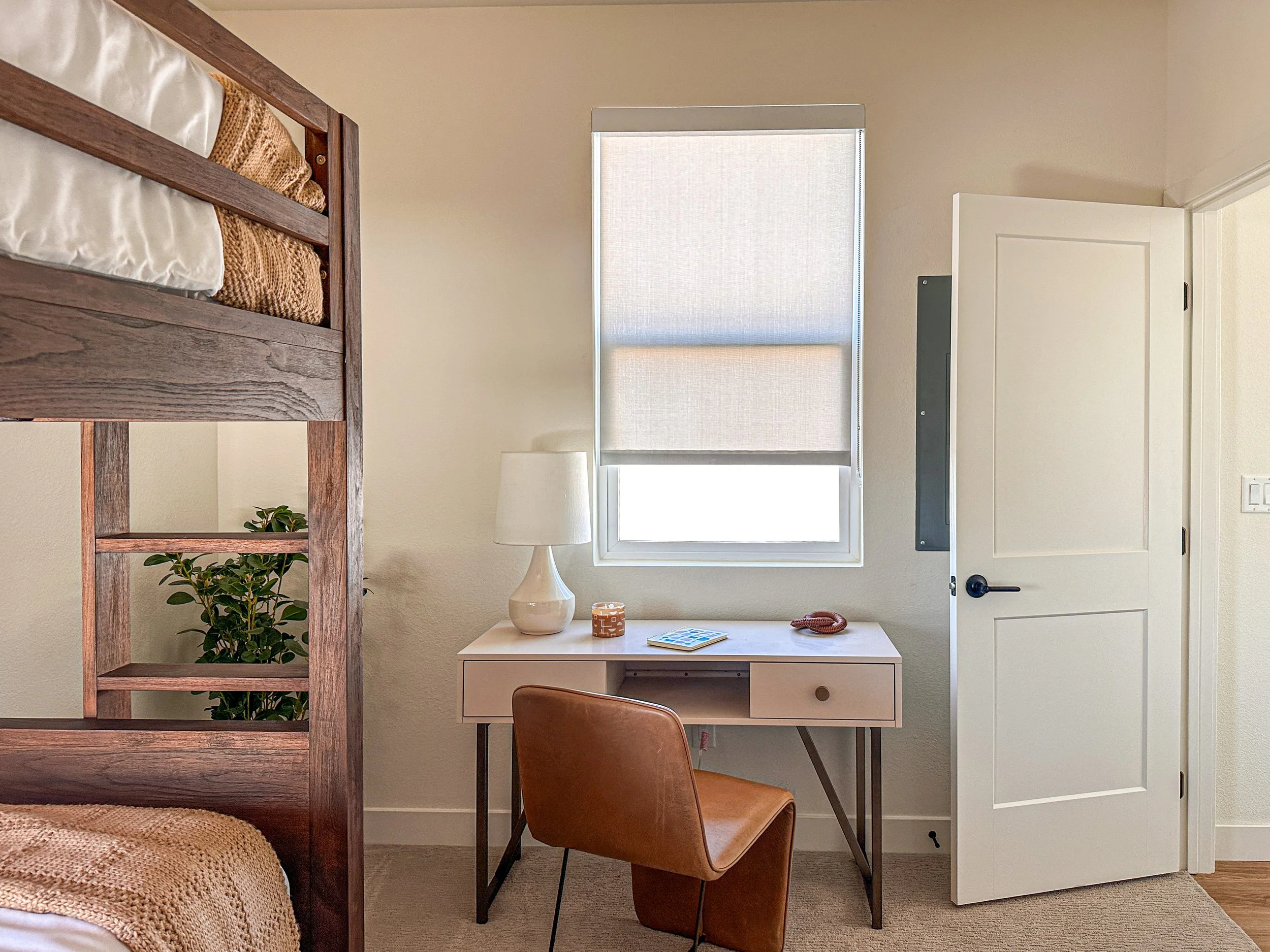 A cozy bedroom corner with a loft bed, a small desk, a brown leather chair, a window with a roller shade, and a door, with a lamp, a book, a small plant, and decorative items on the desk.