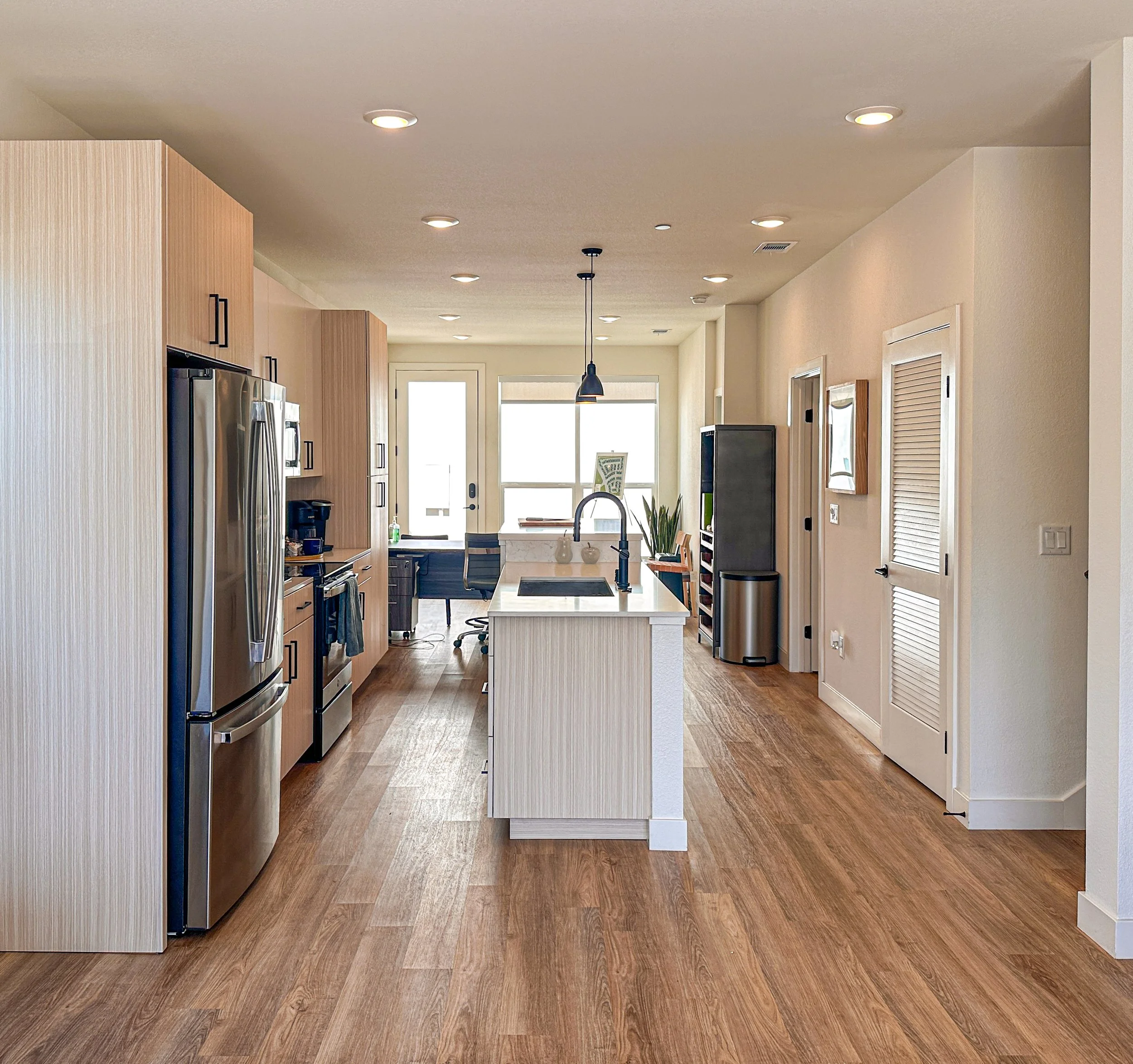 Open-concept kitchen with light wood cabinets, stainless steel refrigerator, white kitchen island, and hardwood floors, with a dining area and sunny windows in the background.
