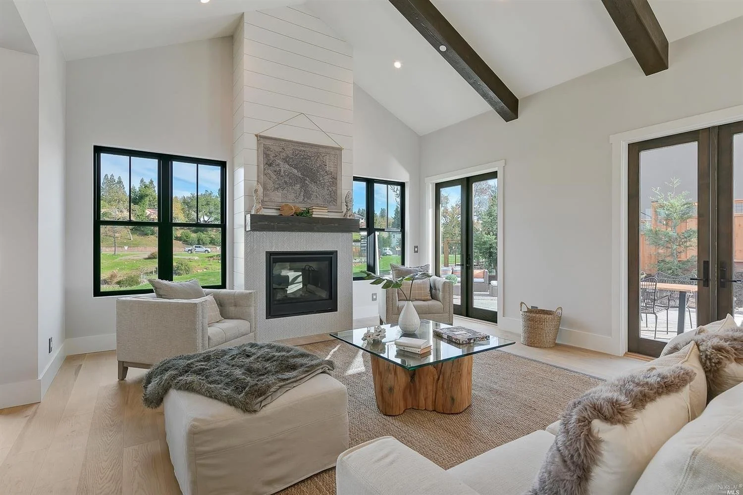 A bright living room with large windows, a fireplace, light-colored sofa, armchair, and a glass coffee table, decorated in a modern rustic style.