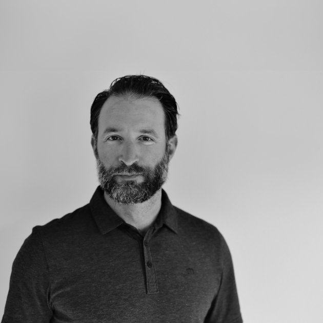 Black and white portrait of a man with a beard and dark hair, wearing a dark collared shirt, standing against a plain light background.