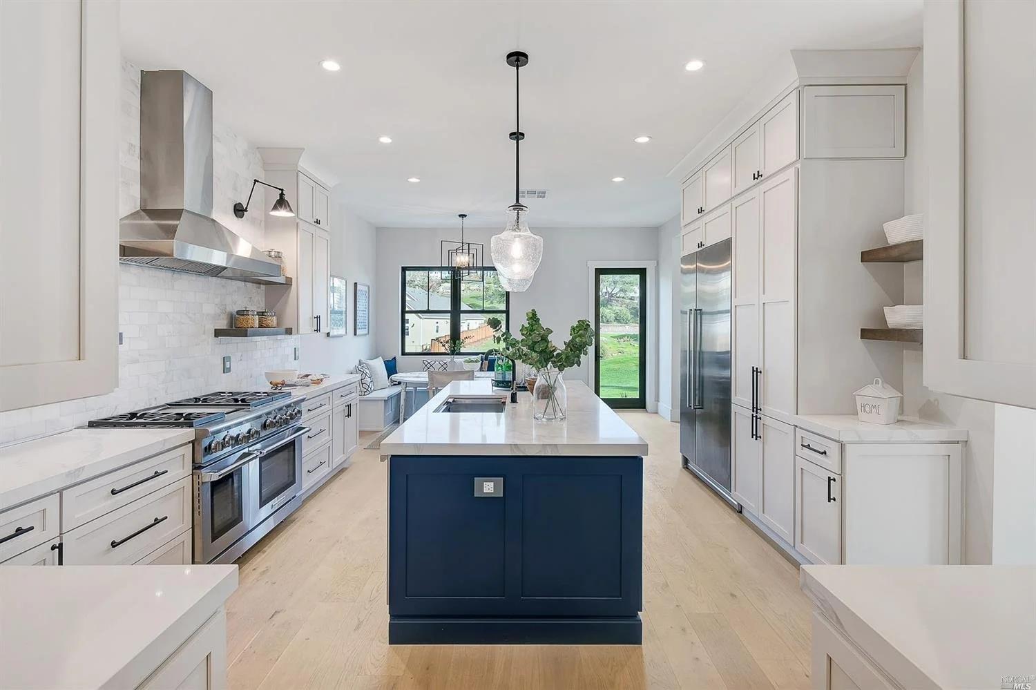 Modern kitchen with white cabinetry, stainless steel appliances, a central island with a dark blue base, and natural light from large windows and glass door.