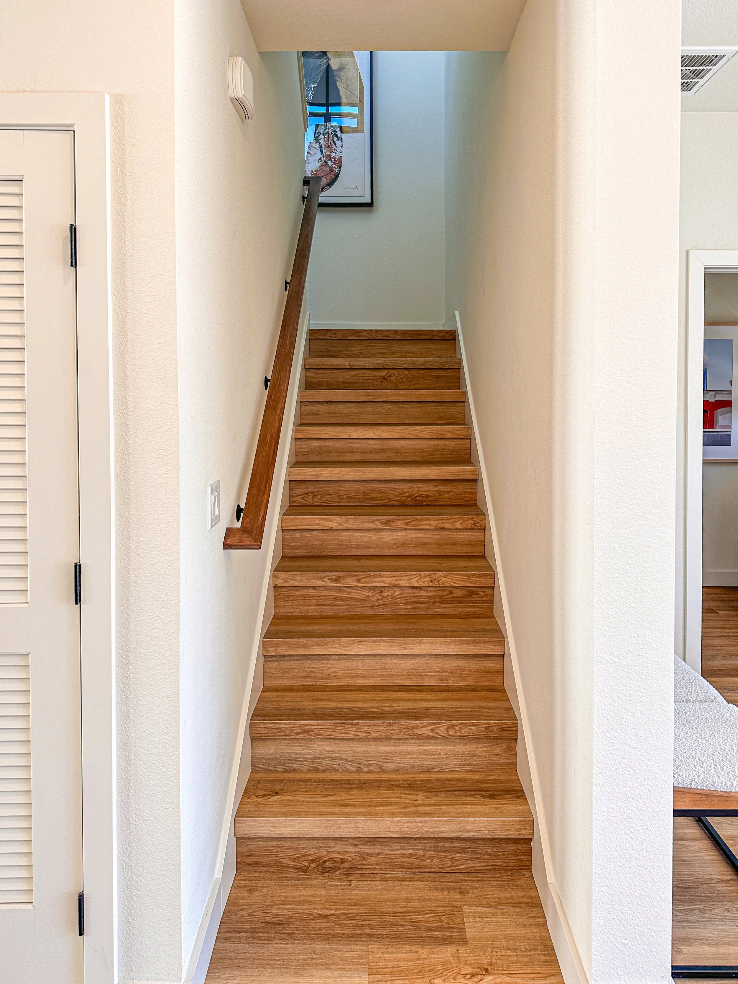 Wooden staircase in a narrow hallway with white walls, a handrail on the left, and artwork at the top of the stairs.