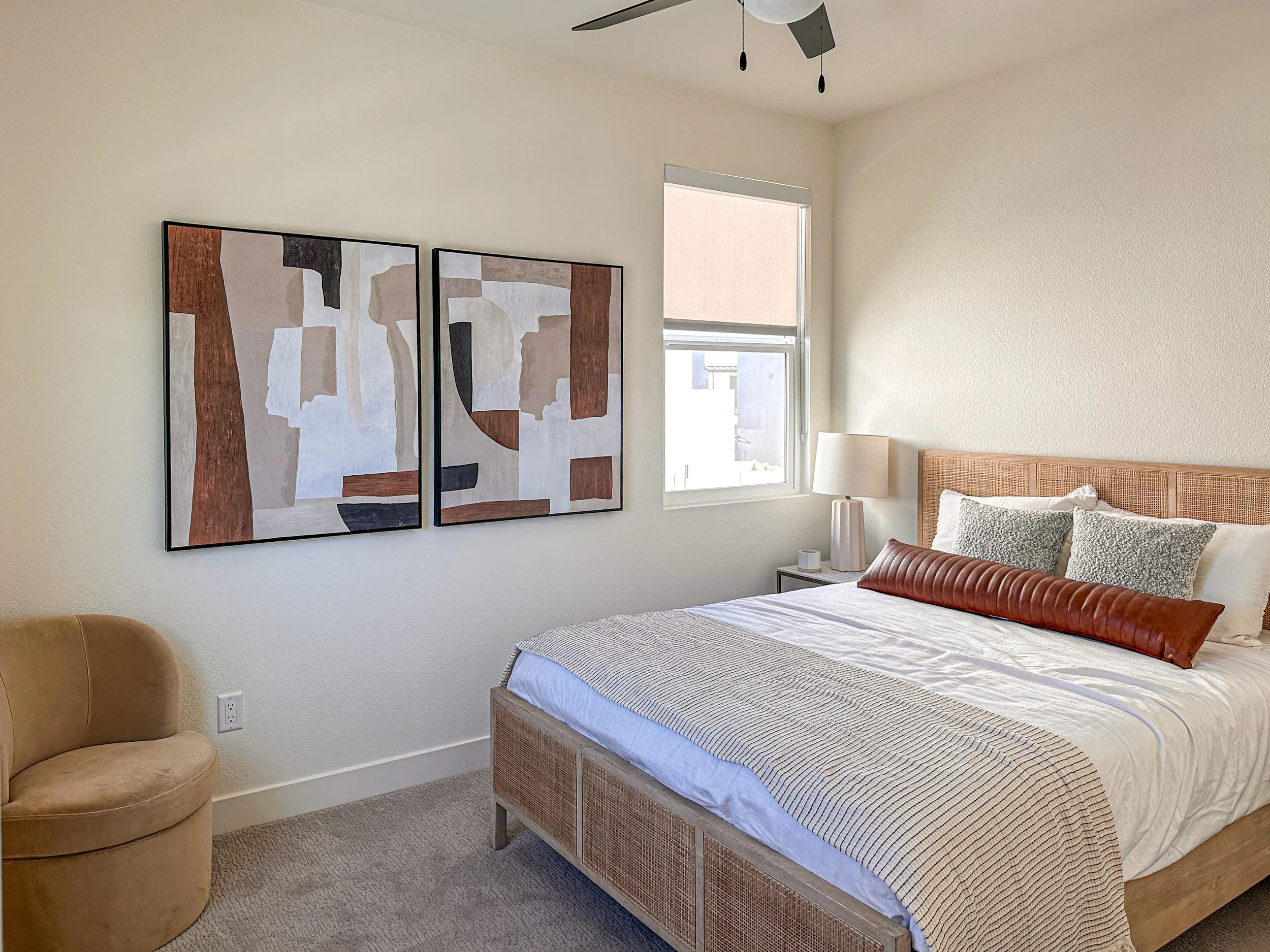 A bedroom with a large bed that has a woven headboard, white and patterned pillows, and a leather bolster pillow. There is a bedside table with a lamp and small decorative items, a window with a roller shade, and abstract art on the wall. A tan uphol
