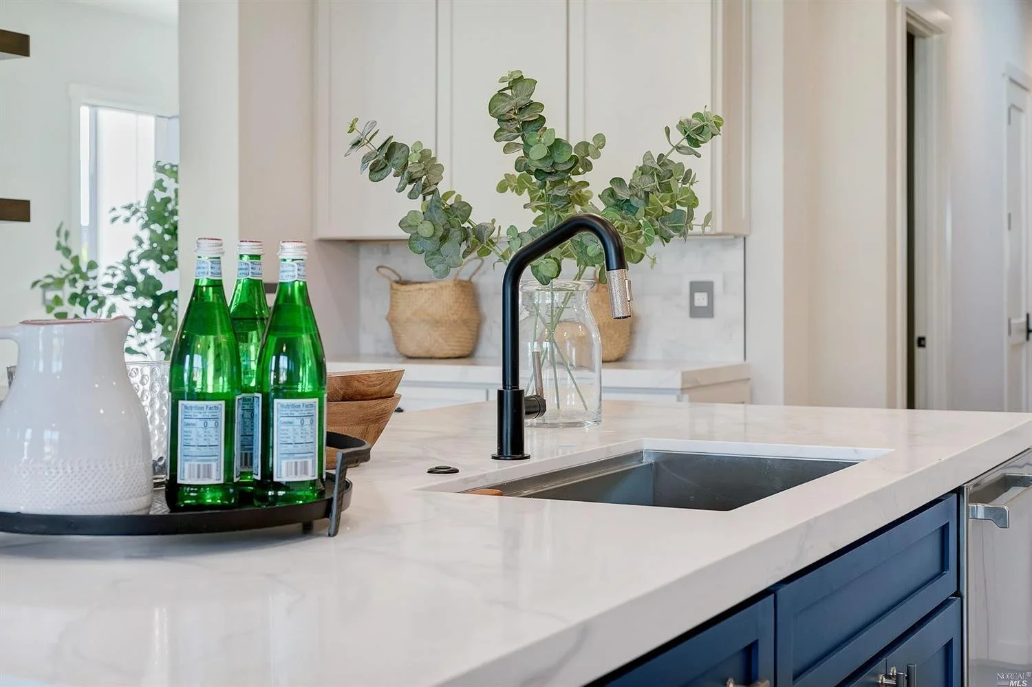 A modern kitchen island with a white marble countertop, black sink, and black faucet. On the island are three green glass bottles, a white pitcher, a wooden bowl, and a large clear glass vase with green eucalyptus branches. The background features white cabinets and decorative baskets.