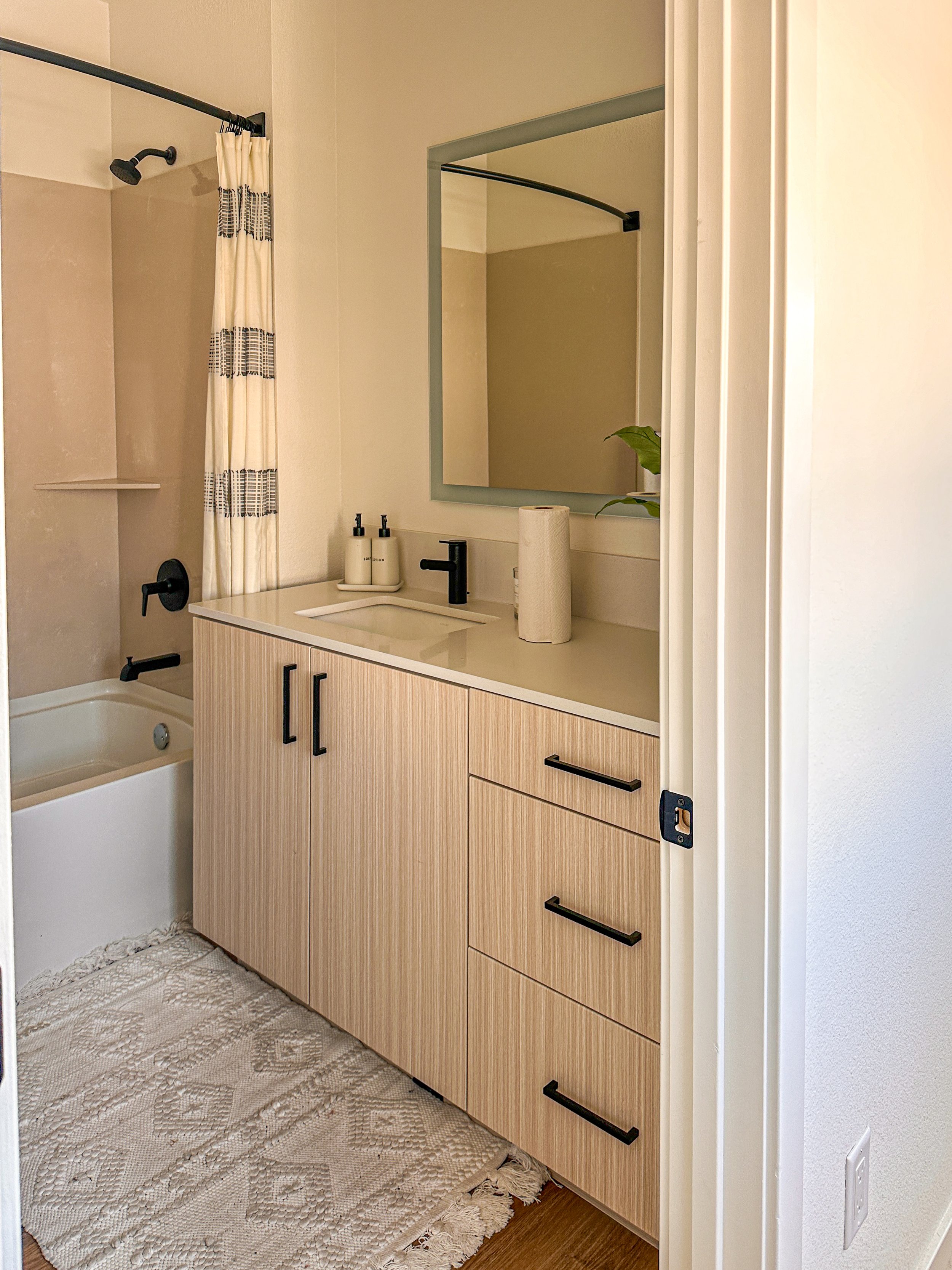 Bathroom with a vanity with a mirror, a sink, black faucet, and black handles on the cabinets. A towel paper holder, hand soap, and a paper towel roll are on the vanity. There is a bathtub with a shower curtain. A rug is on the wooden floor.