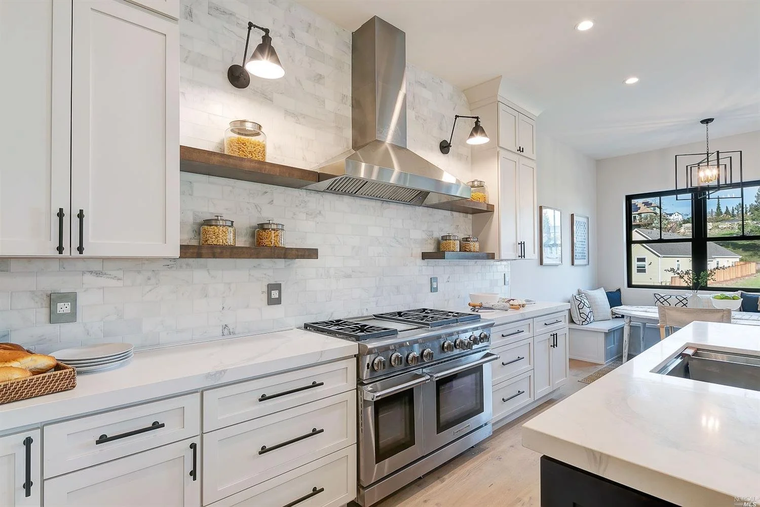 Modern kitchen with white cabinets, stainless steel stove, white marble backsplash, and floating wooden shelves with jars of pasta. Natural light from large window, dining nook with bench and pillows nearby.