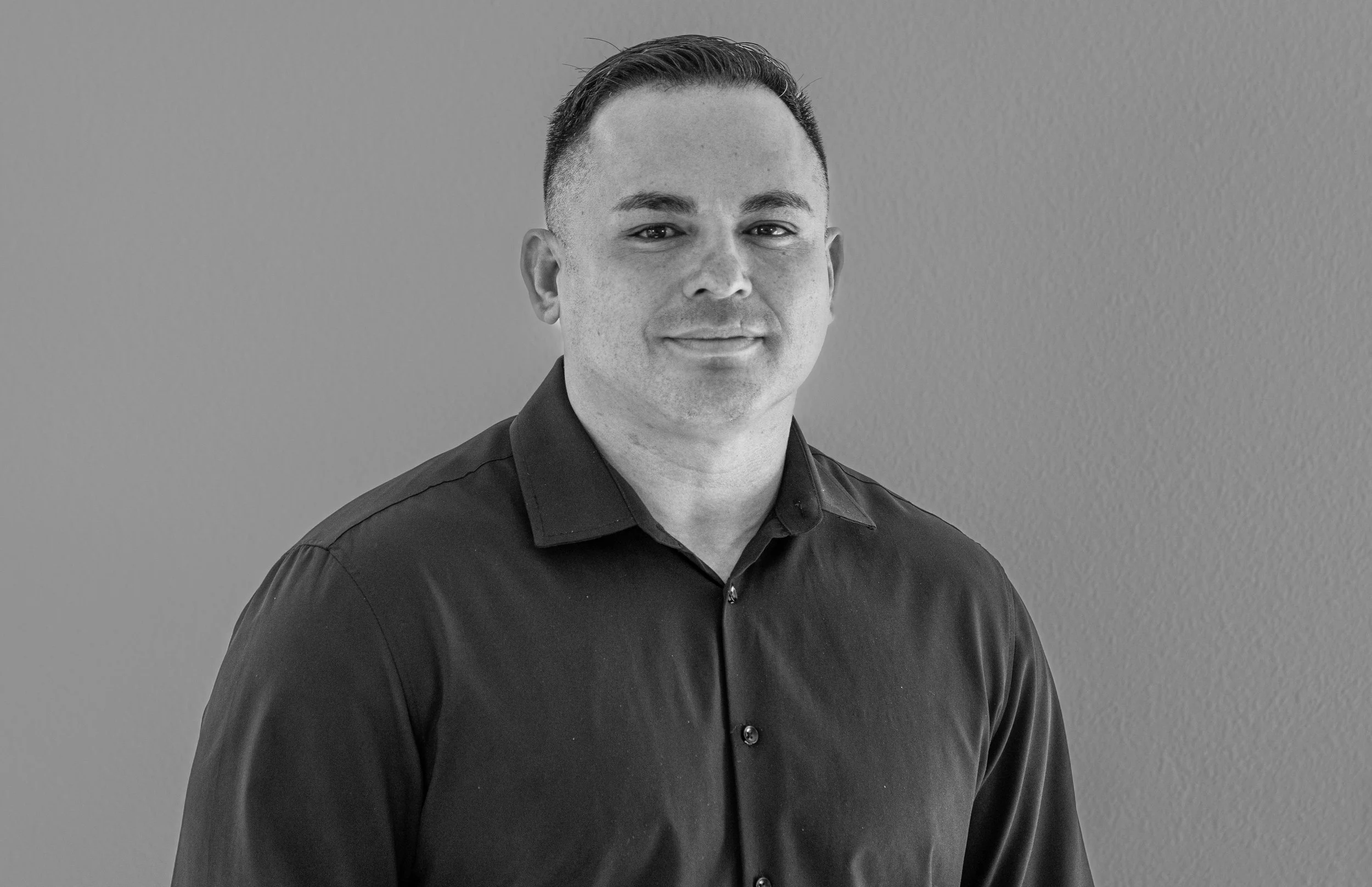 Black and white photo of a man in a dark collared shirt standing against a plain wall.