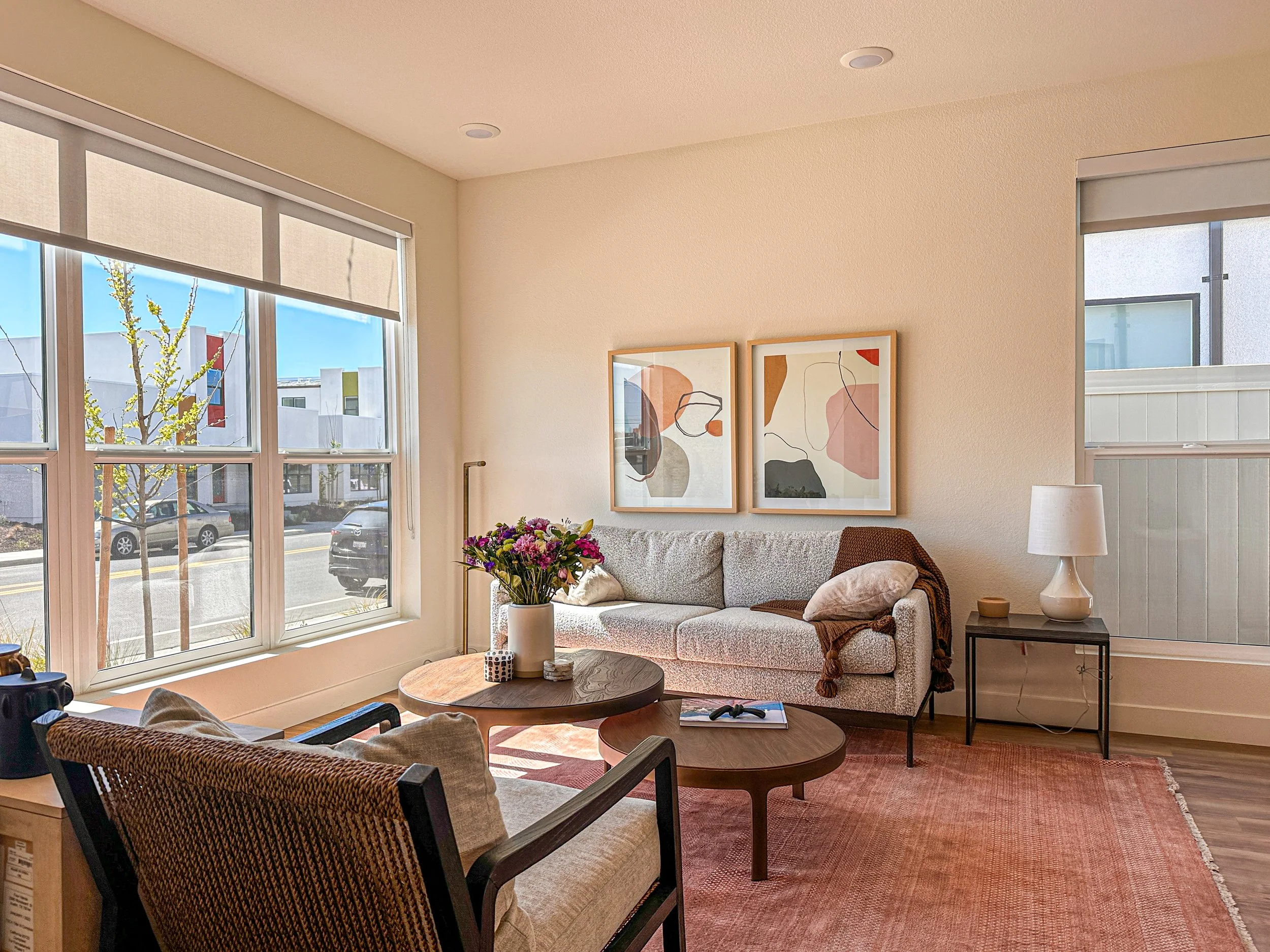 Bright living room with large windows, a beige sofa with pillows, two abstract art pieces, a coffee table with a vase of flowers, and a side table with a lamp.