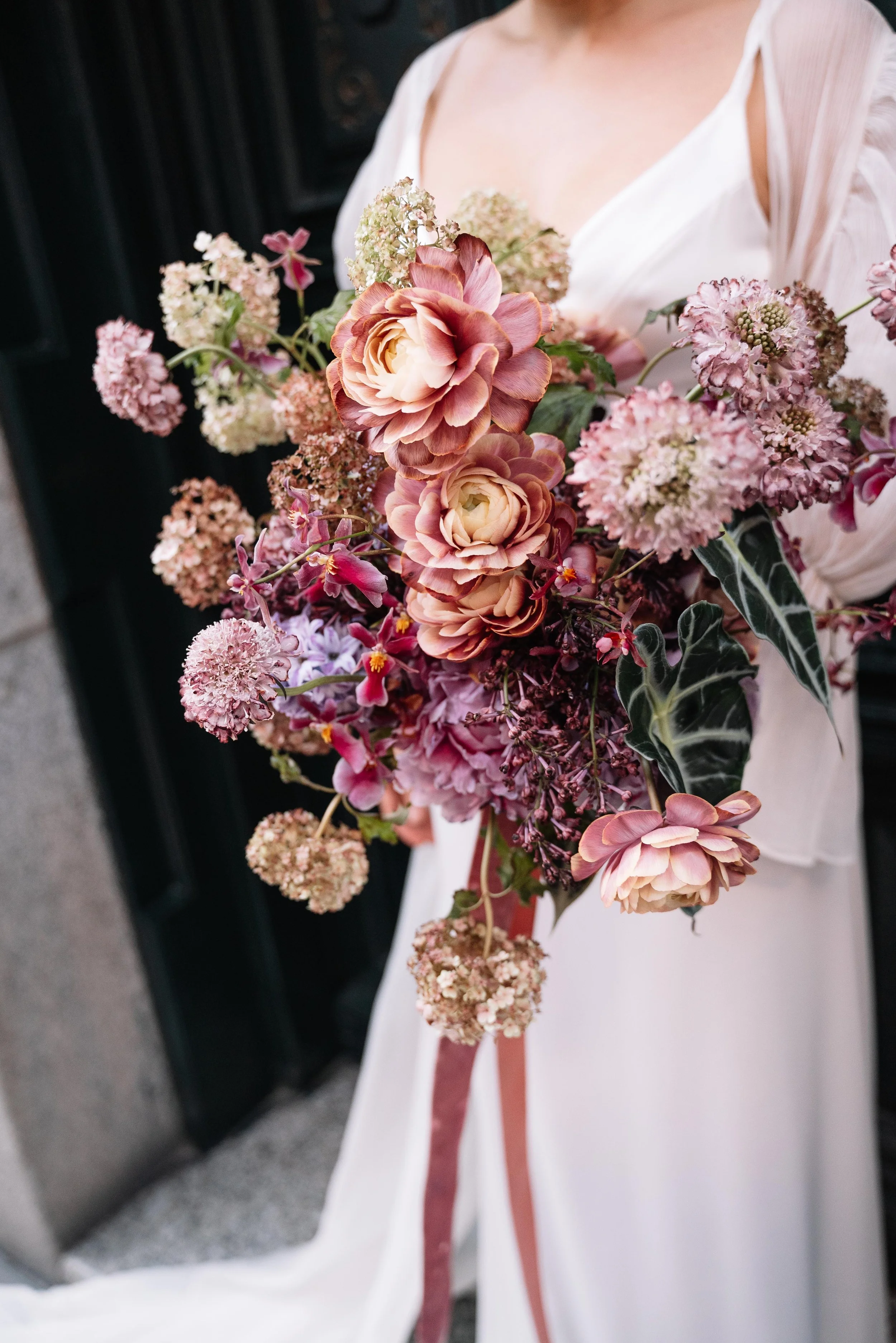 Ramo de flores con diversas flores en tonos rosados y verdes, llevado por una persona con vestido blanco, parcialmente visible.