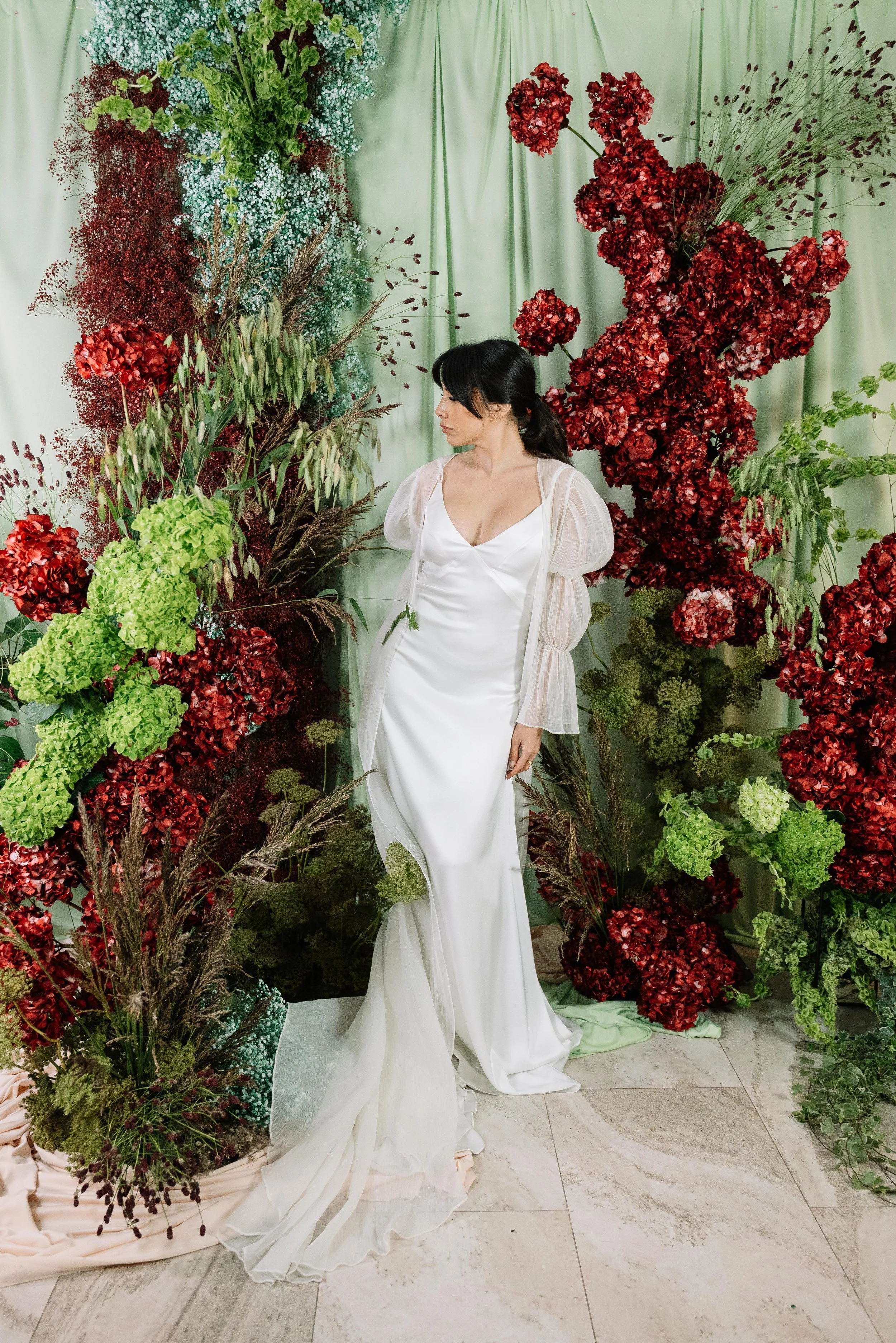 Mujer vestida con vestido blanco en un entorno de flores rojas, verdes y moradas, con cortinas verdes en el fondo.