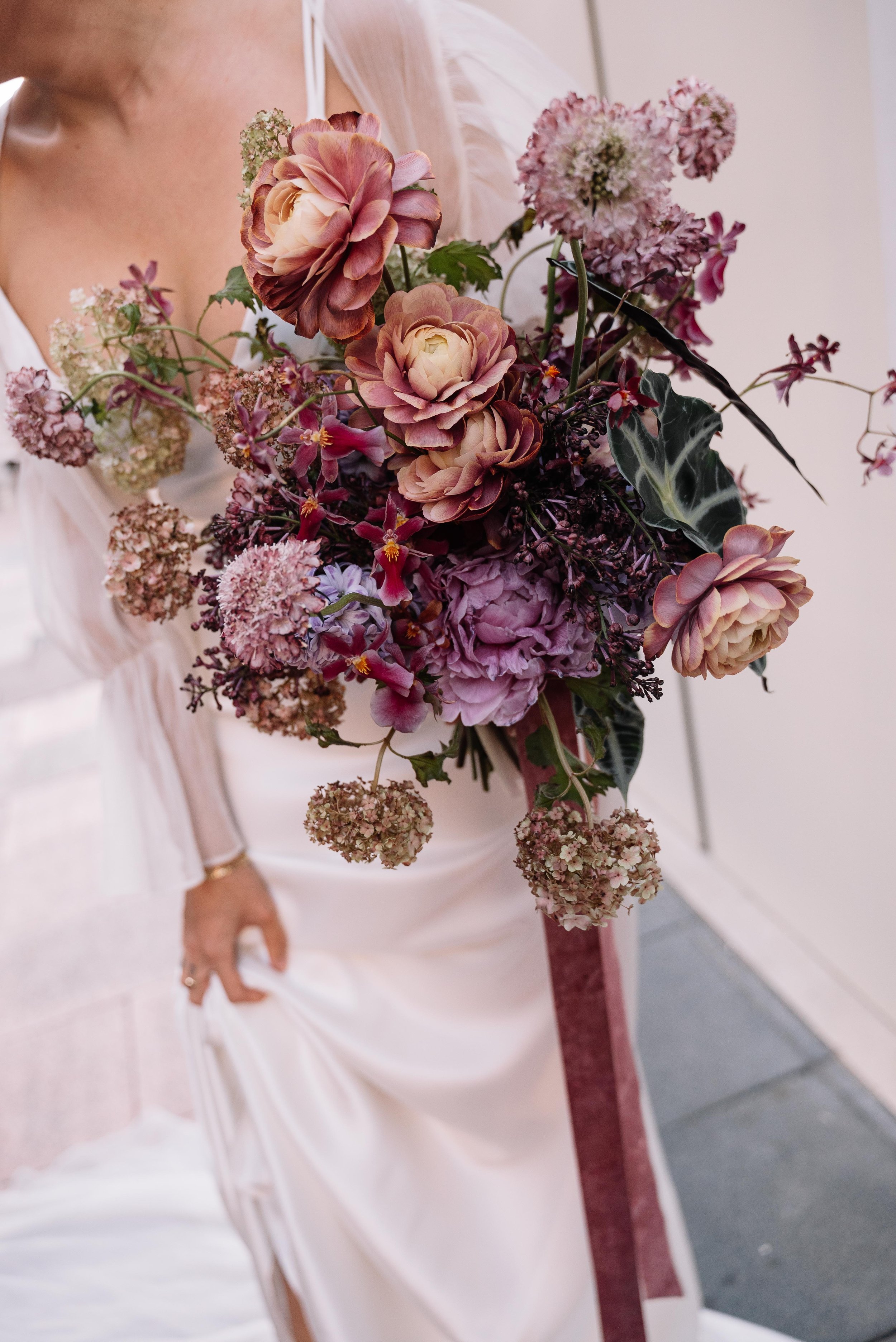 Persona sosteniendo un ramo de flores en tonos rosados y lavanda, con vestido blanco.