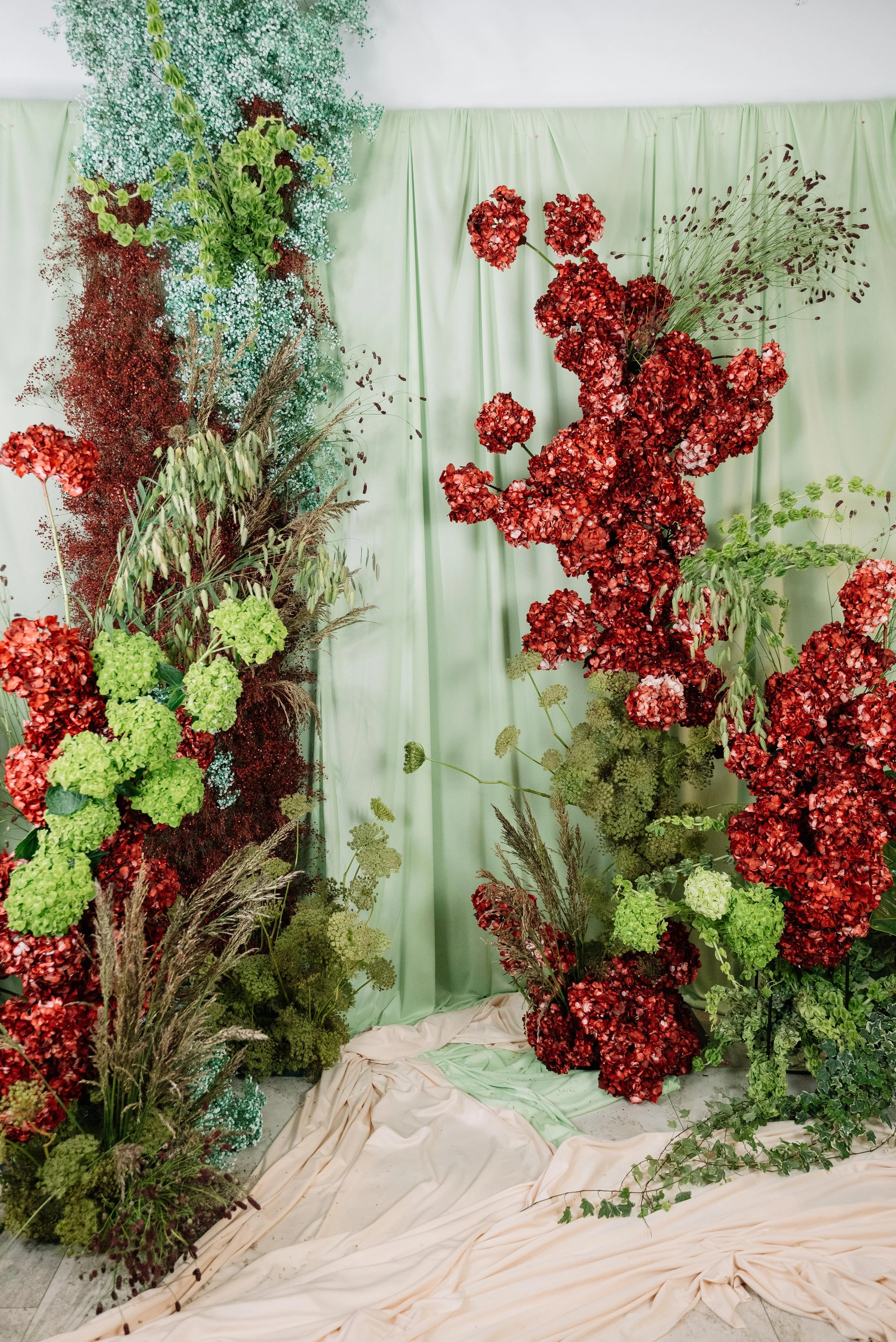 Decoración con flores y plantas en tonos rojos y verdes con cortinas verdes de fondo y tela beige en el suelo.