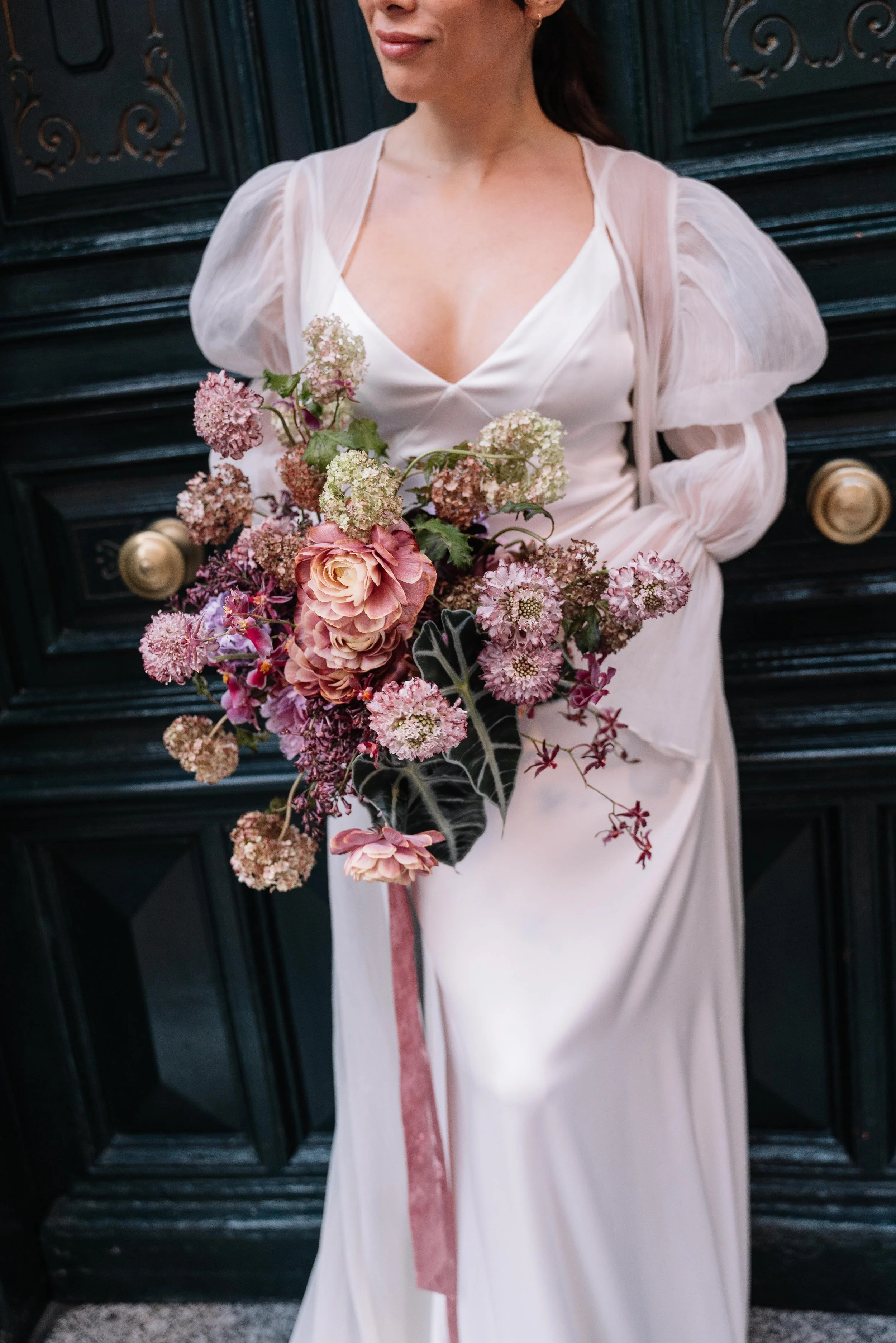 Una mujer con vestido blanco y mangas de tul sostiene un ramo de flores en tonos rosa, blanco y gris, frente a una puerta oscura con detalles dorados.