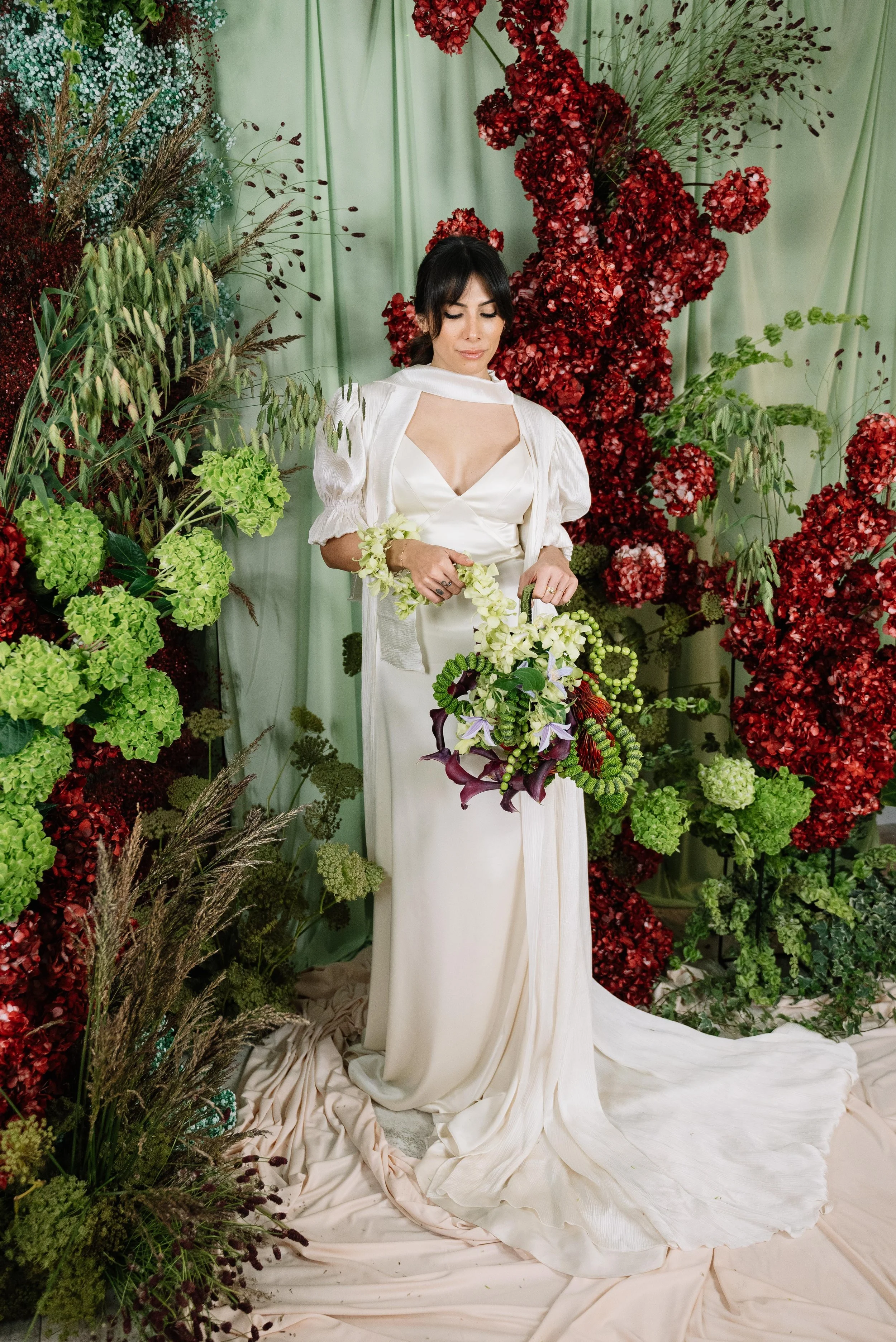 Mujer en vestido blanco y cabello oscuro, rodeada de flores verdes y rojas, sostiene un ramo de flores blancas y moradas en un escenario decorado con cortinas verdes y plantas.