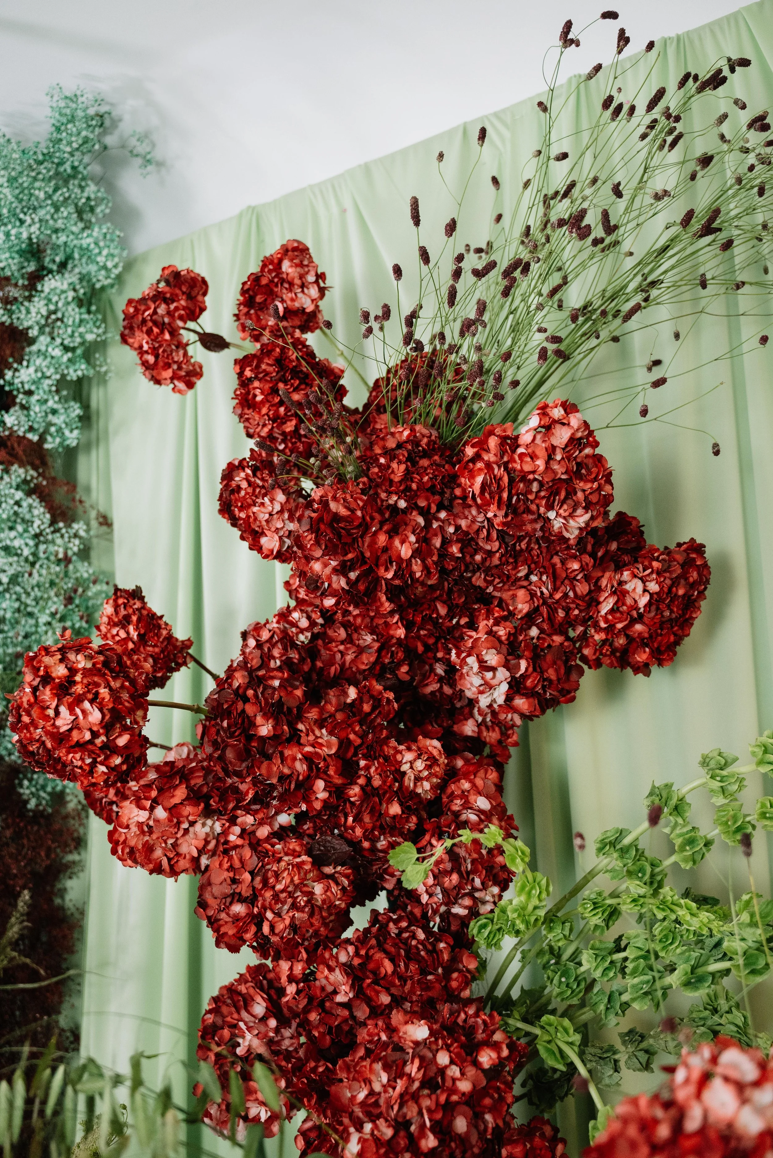 arreglo floral con grandes racimos de flores rojas y plantas verdes, sobre fondo de cortina de color menta