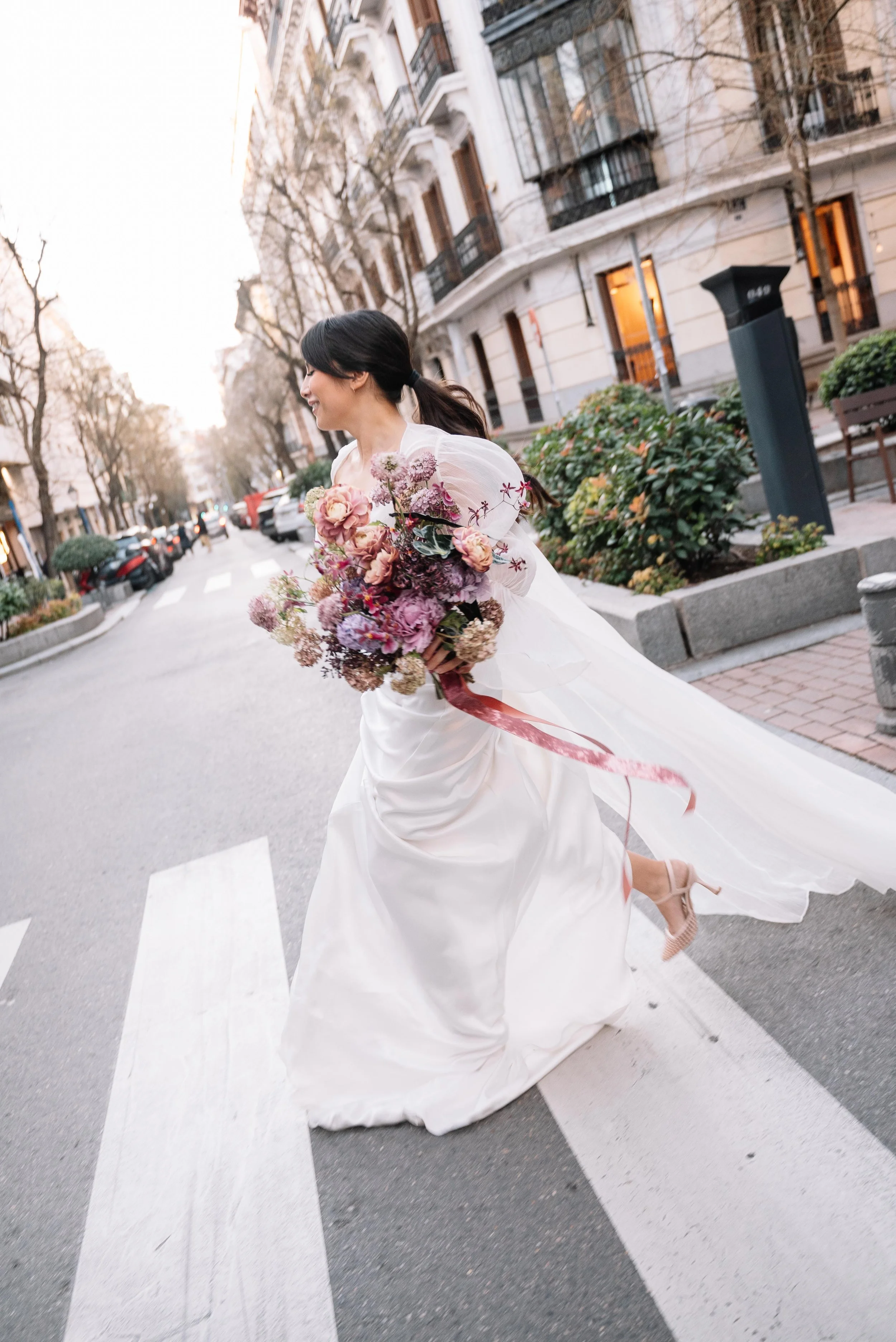 Una mujer con vestido de novia sonriente cruzando una calle con flores en las manos durante el amanecer en una calle urbana