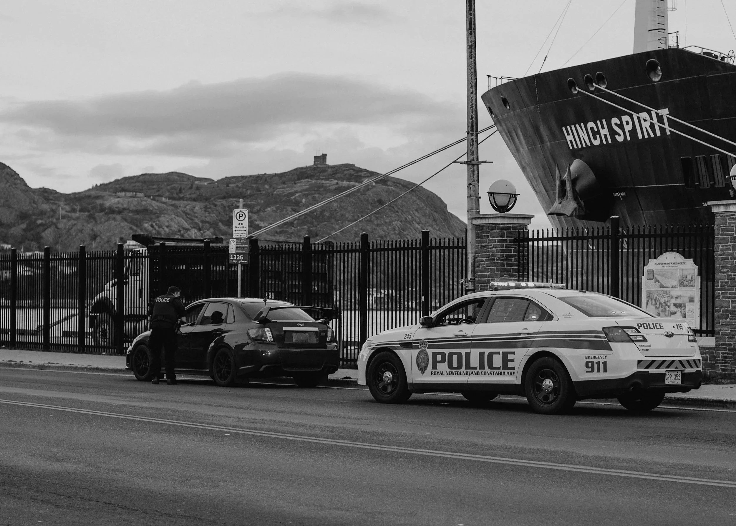 Police officer speaking to the driver of a black car while standing on the sidewalk. Two police cars marked with 'POLICE 911' and 'ROYAL NEWFOUNDLAND AND LABRADOR CONSTABULARY' are parked nearby. A large ship with 'Hinch Spirit' on its bow is docked behind a fence, with a scenic hilly landscape and a monument visible in the background.
