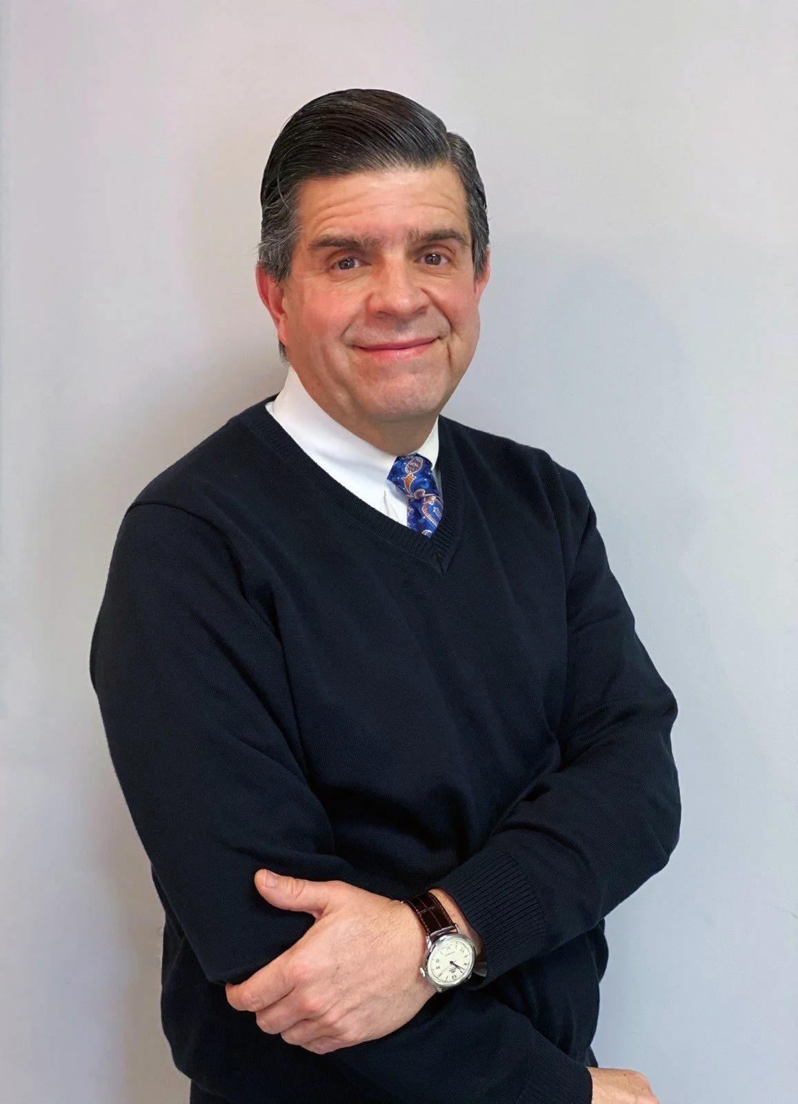 A man with dark slicked-back hair wearing a white dress shirt, a patterned blue tie, and a black V-neck sweater, standing with crossed arms against a white background.