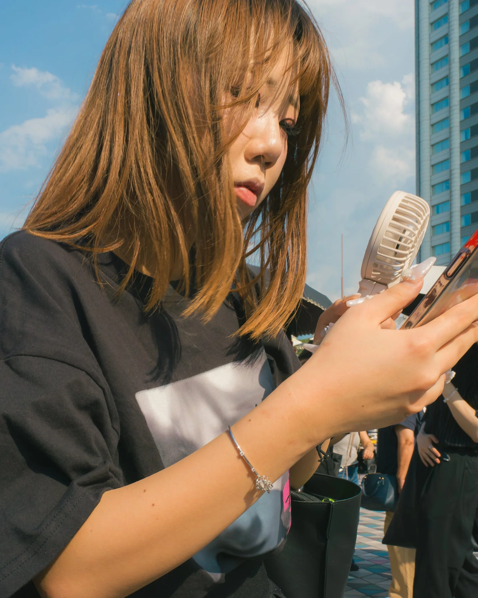 A woman with long hair using her phone outdoors during daytime amidst other people.