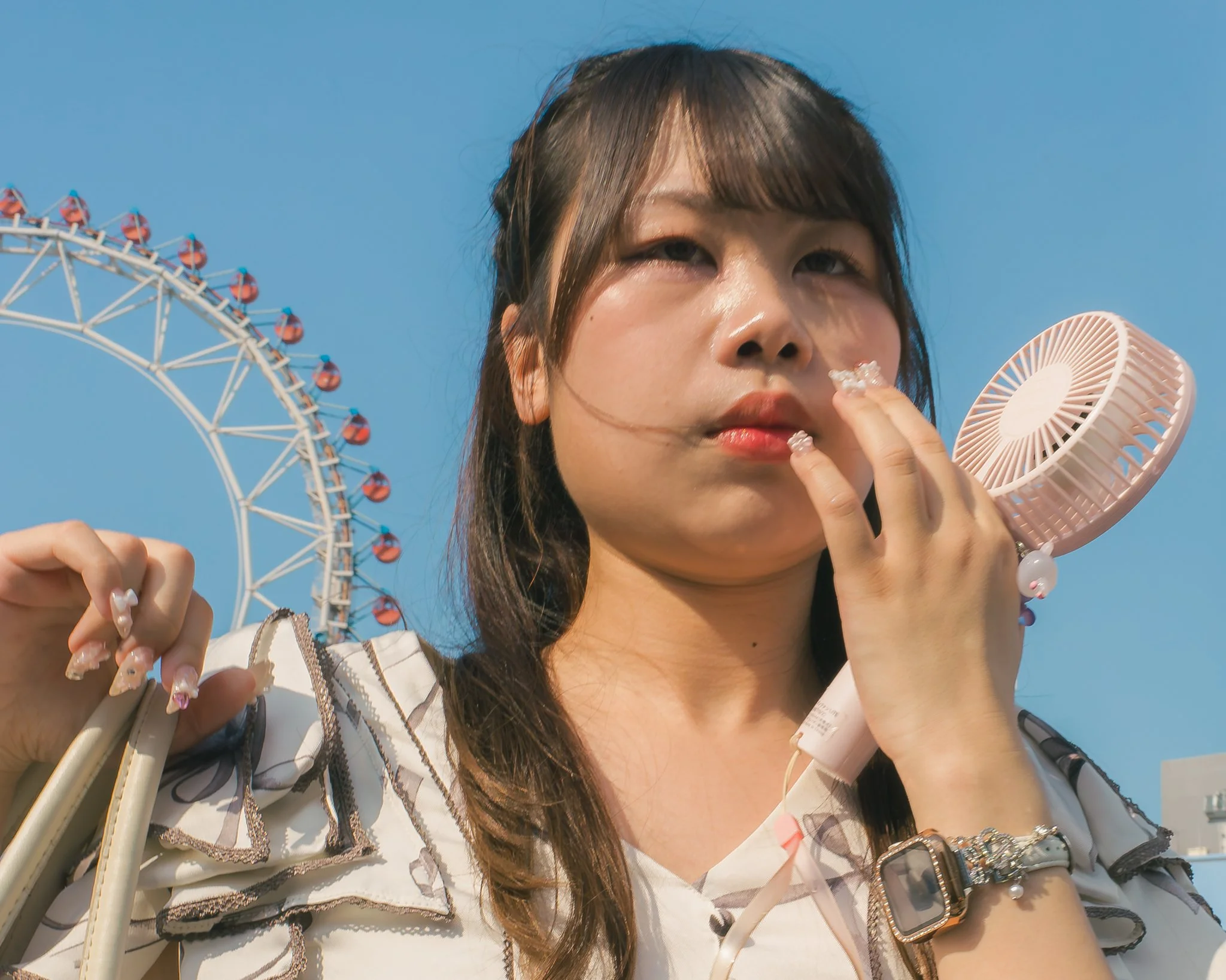 Close-up of a young woman with black hair holding a handheld fan and a cone-shaped object, with a Ferris wheel in the background against a clear blue sky.