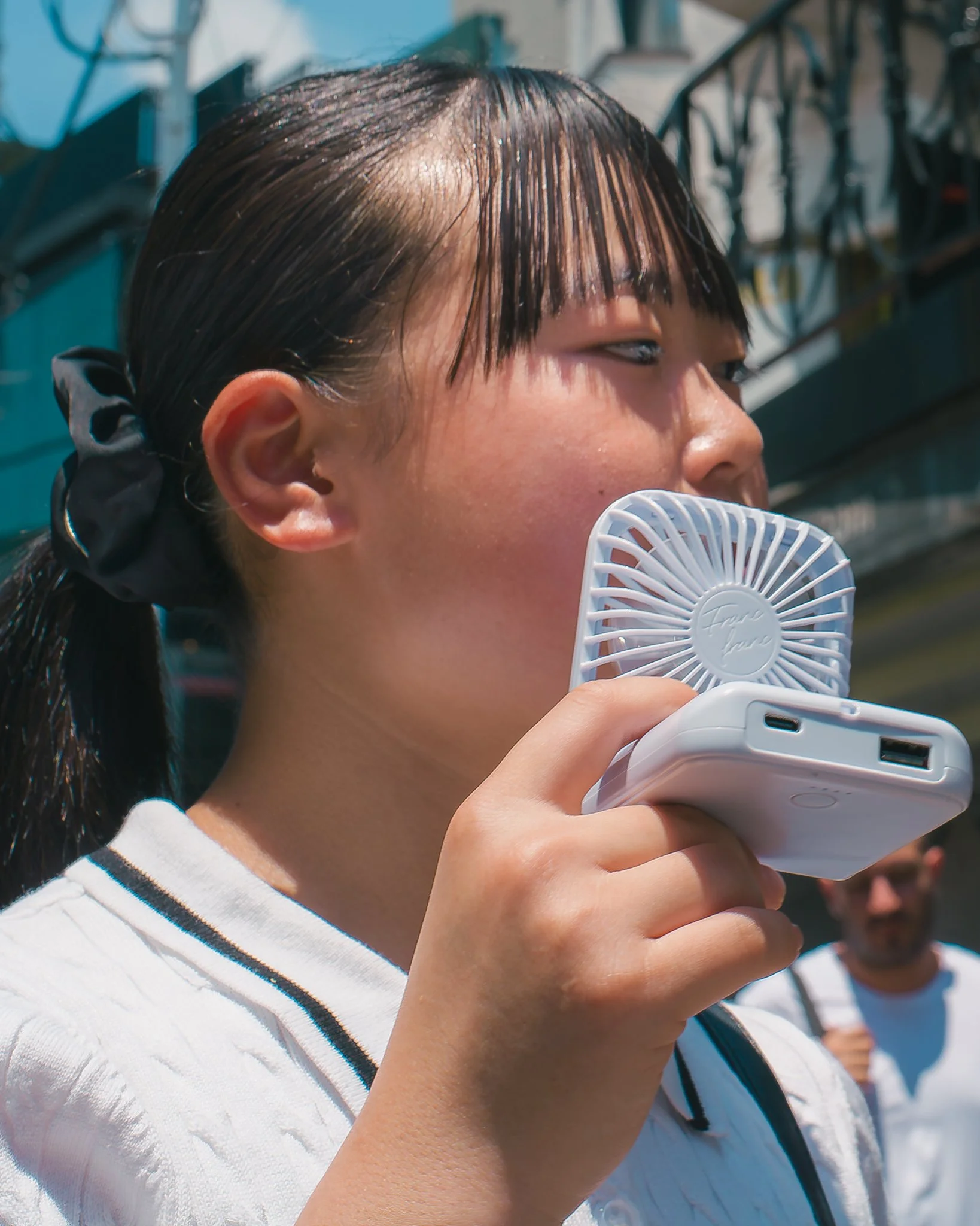 A woman with wet, dark hair holding a small, white handheld fan and a smartphone, standing outdoors on a sunny day.
