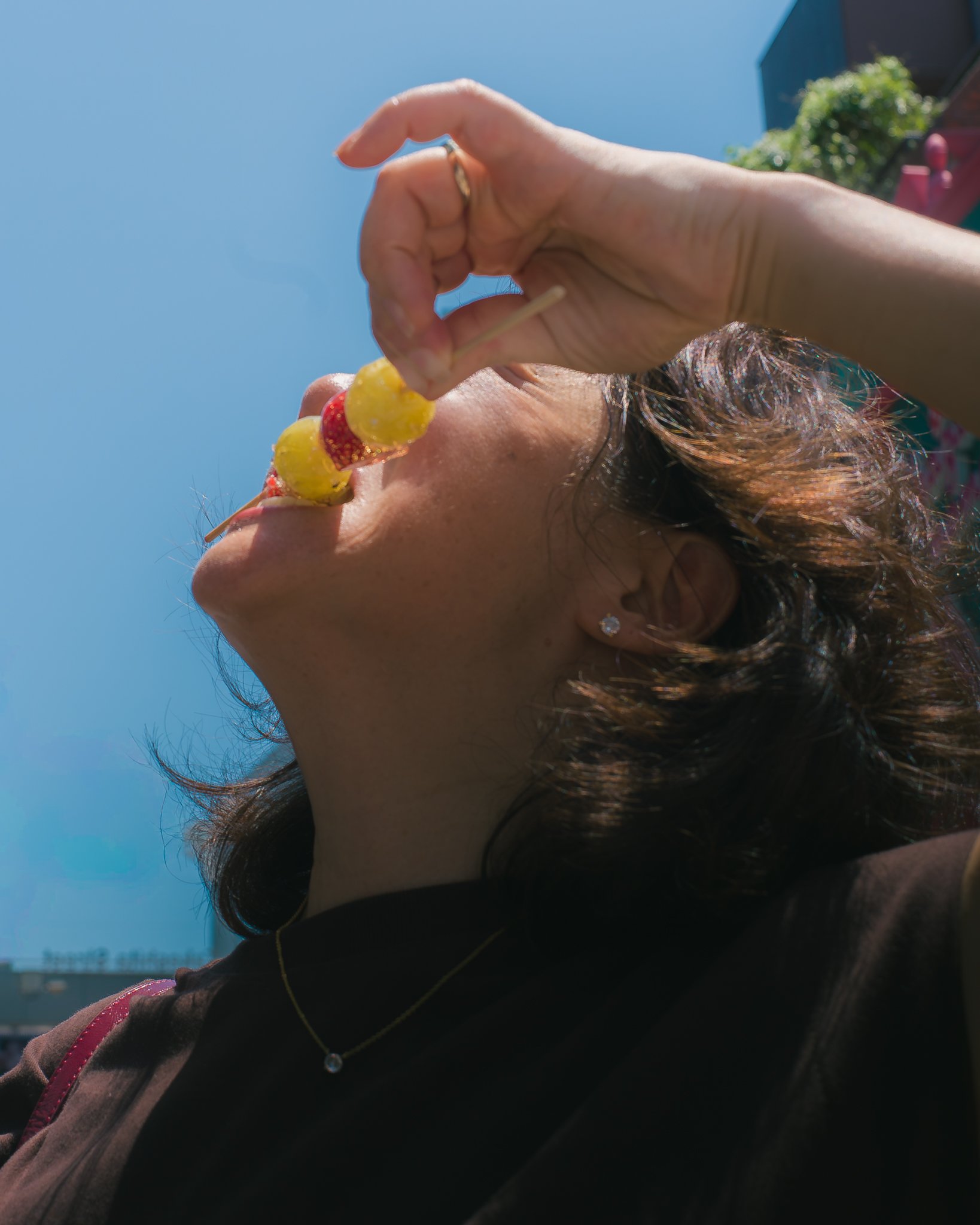 A woman with curly hair, wearing earrings, a necklace, and a dark shirt, is holding a spiral-shaped lollipop with yellow and red colors up to her mouth against a clear blue sky background.