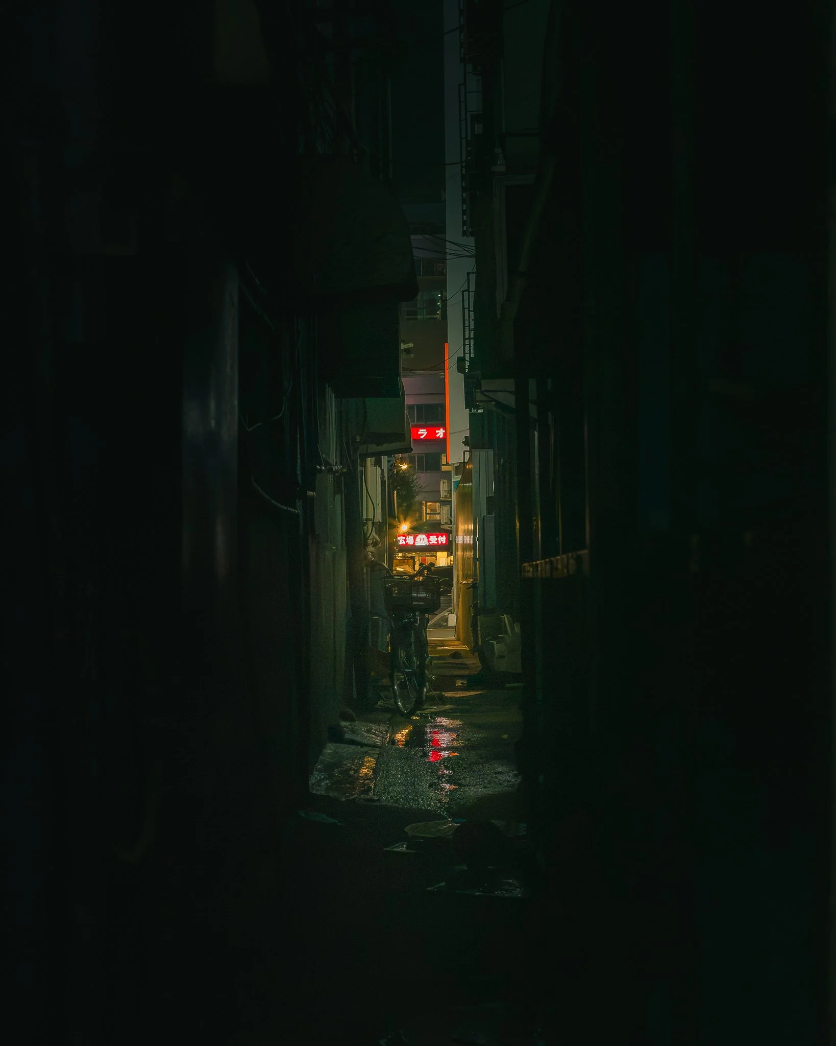 Nighttime image of a city street viewed from inside a building with a wet floor. The scene is dark, illuminated by a few streetlights and glowing signs, including a red and white sign with Japanese characters. Several parked cars are visible along th
