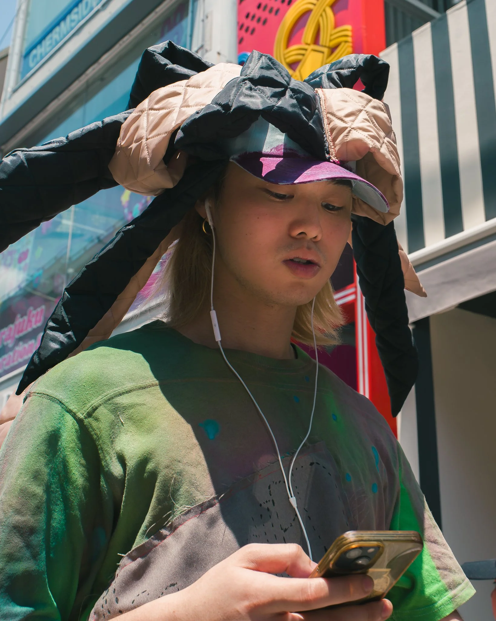 A young woman with blonde hair wearing a tie-dye t-shirt, listening to earphones, and holding a phone while standing outdoors in front of colorful signage.