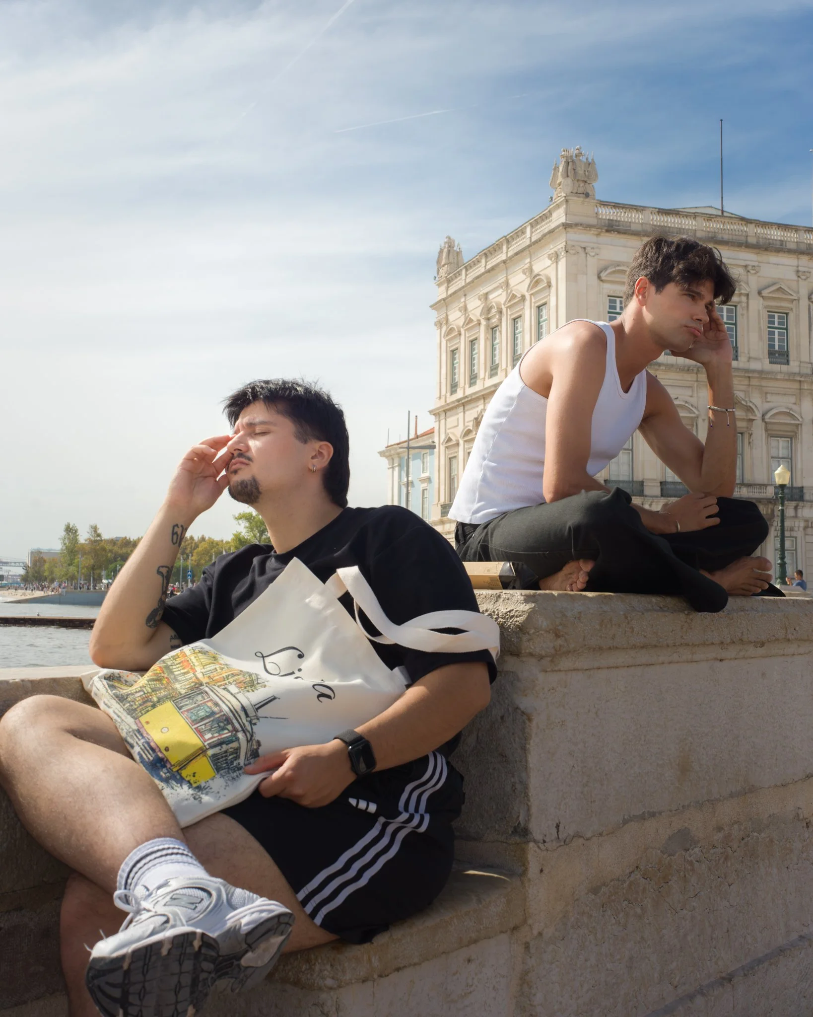 Two young men sit on a stone ledge outdoors near a historical building with classical architecture. Both are engaged in a meditative or relaxing pose, with eyes closed and hands positioned near their foreheads, in a peaceful setting on a sunny day.