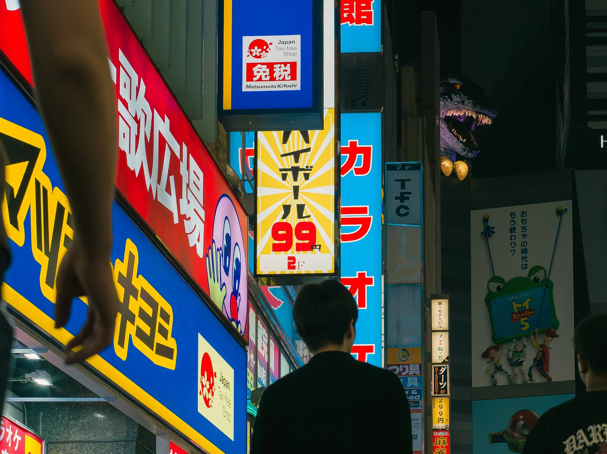 Night scene of a busy street with bright, colorful advertisements and signs in Japanese, including a large blue sign for a tax-free shop, cartoon characters, and a dinosaur figure illuminated in the background. Several pedestrians are visible.
