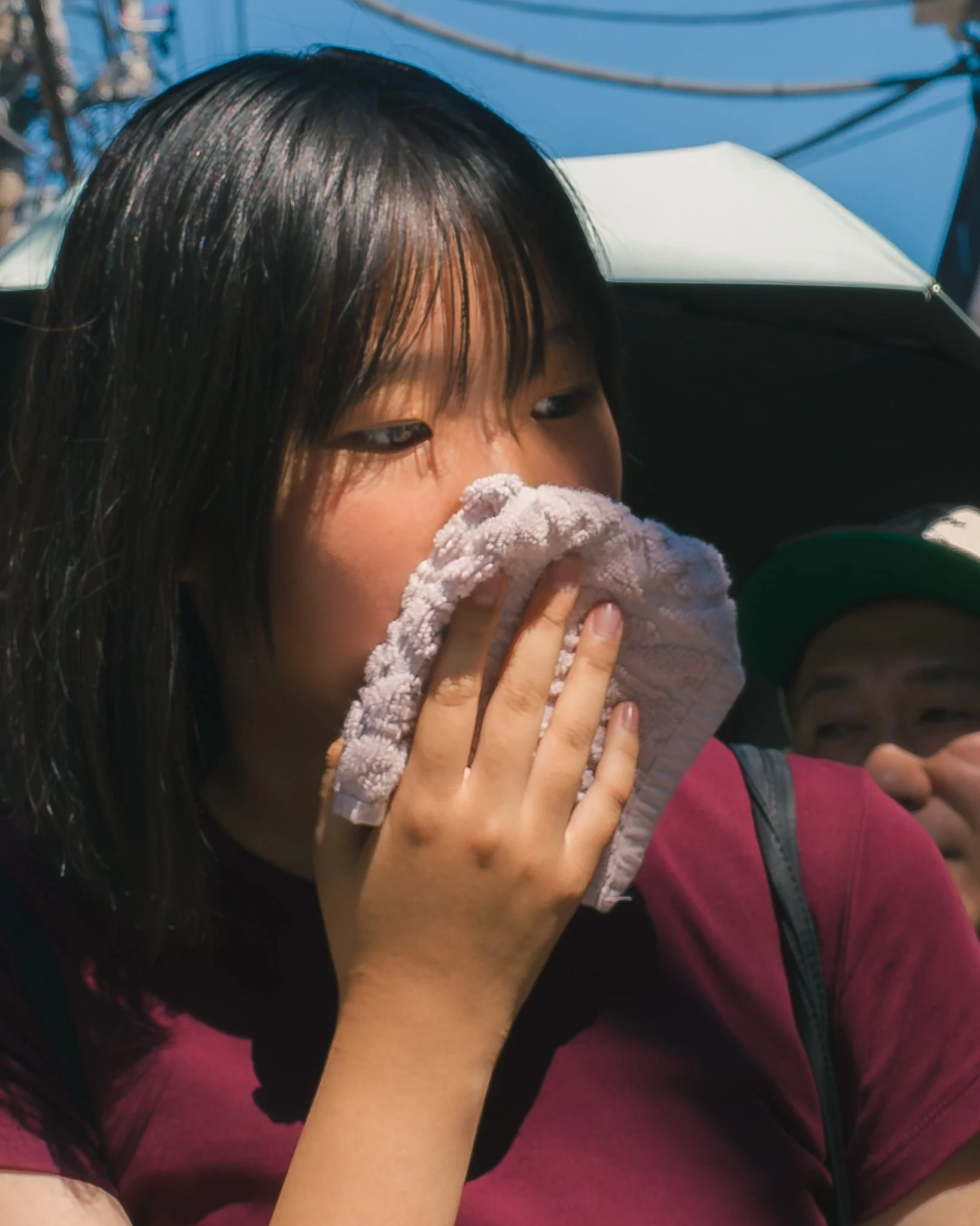 A woman with straight black hair is partially covering her face with a pink towel or cloth, wearing a dark-colored shirt, and appears to be outdoors, with a blue sky and some wires in the background.