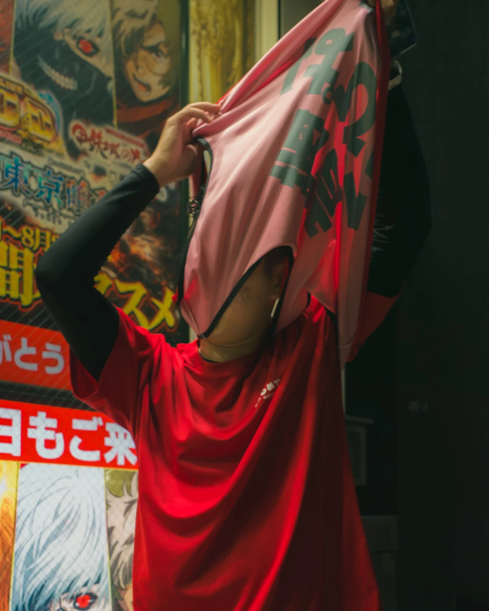 A person in a red long-sleeve shirt is holding up a pink fabric with large dark green printed text in front of a colorful Japanese manga or anime poster.