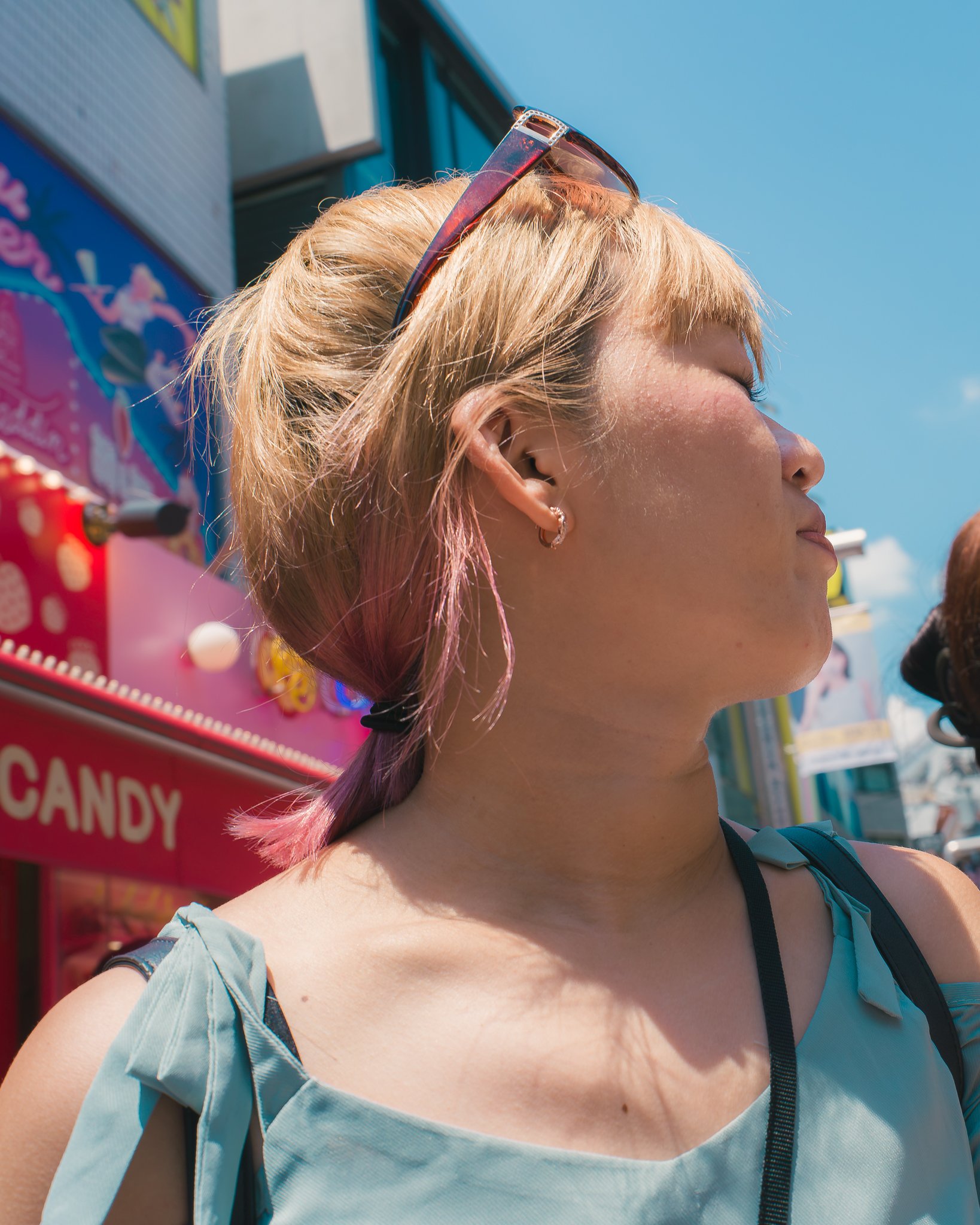 A woman with short, dyed pink hair and sunglasses resting on her head is outdoors on a sunny day, with a colorful candy stand sign in the background.