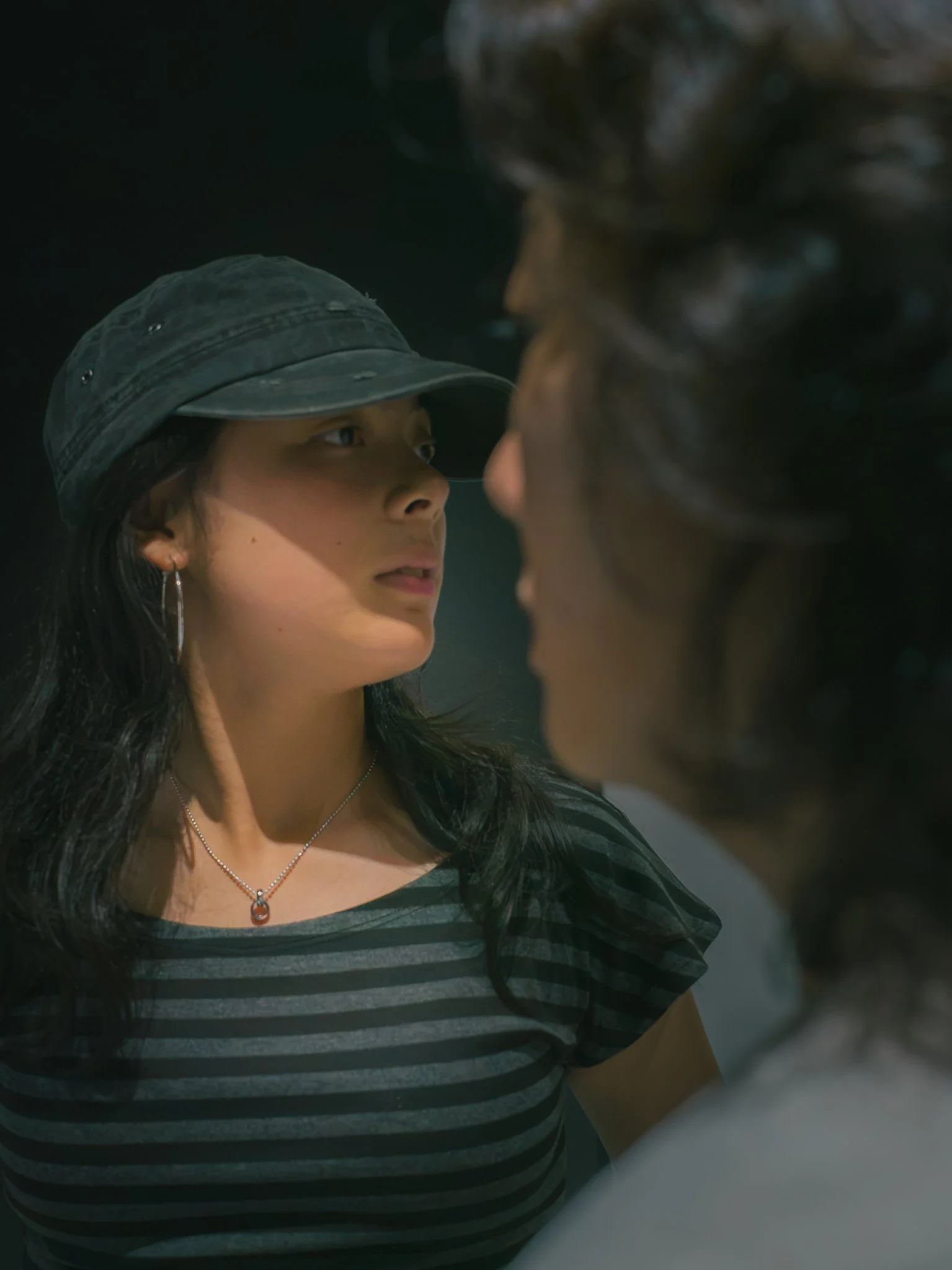 Two women face each other closely, one wearing a bucket hat and the other with curly hair, in a dark setting.