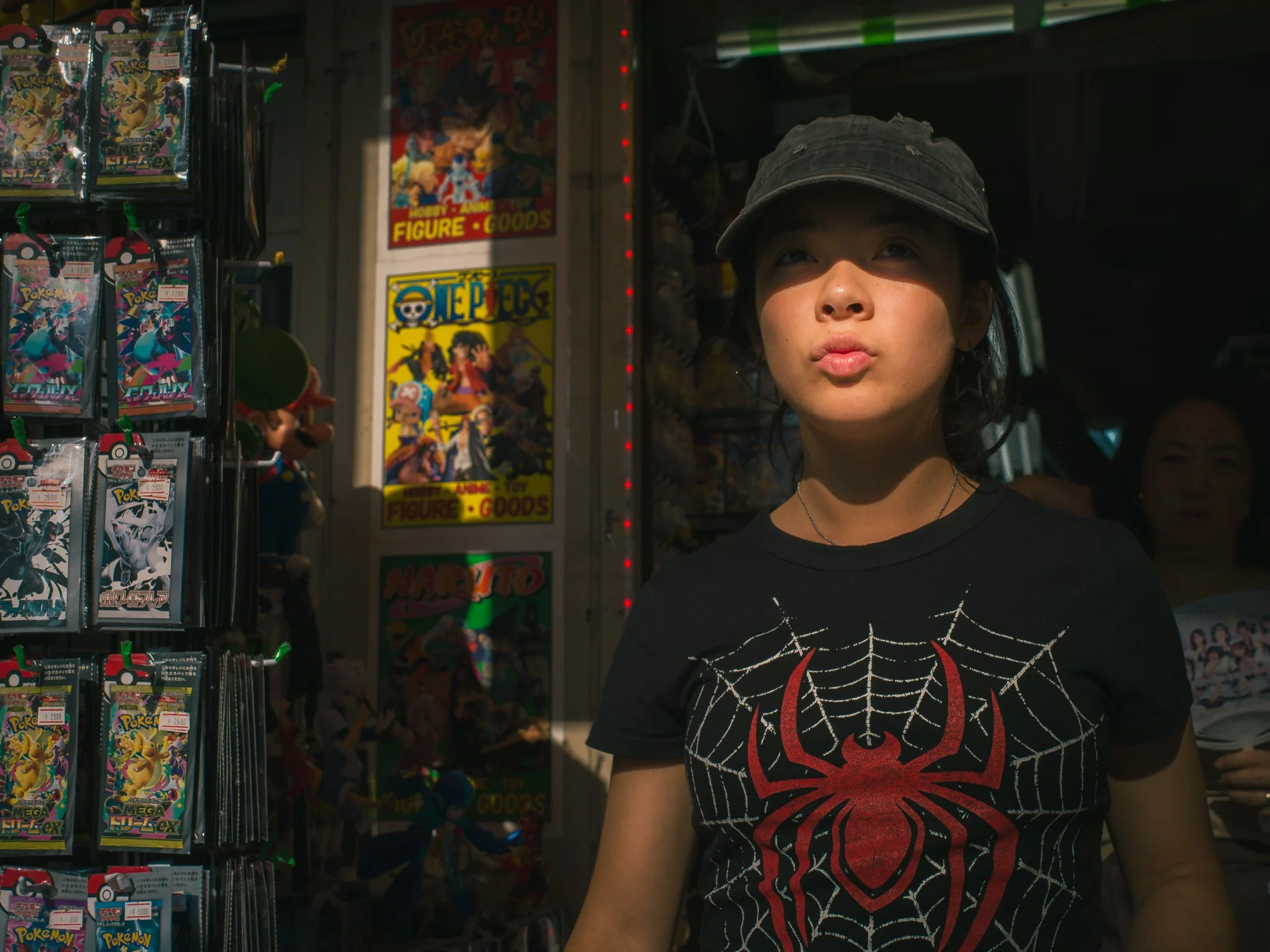 A person wearing a black Spider-Man t-shirt and a black cap standing in a store aisle with Pokémon and One Piece merchandise.