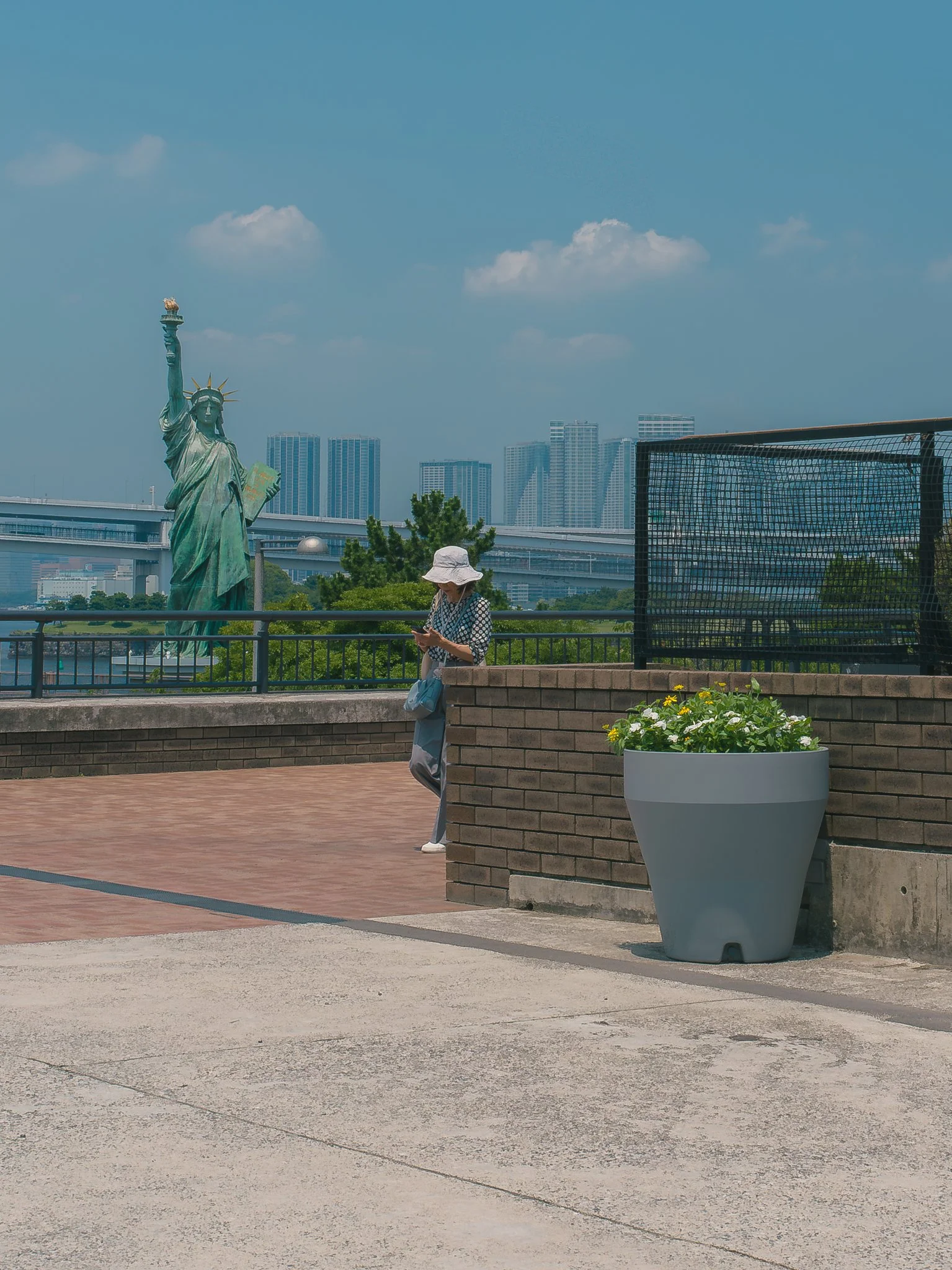 A woman wearing a large white sun hat and polka dot top is walking past a large white planter with yellow and white flowers, near the Statue of Liberty replica and modern skyscrapers in the background.