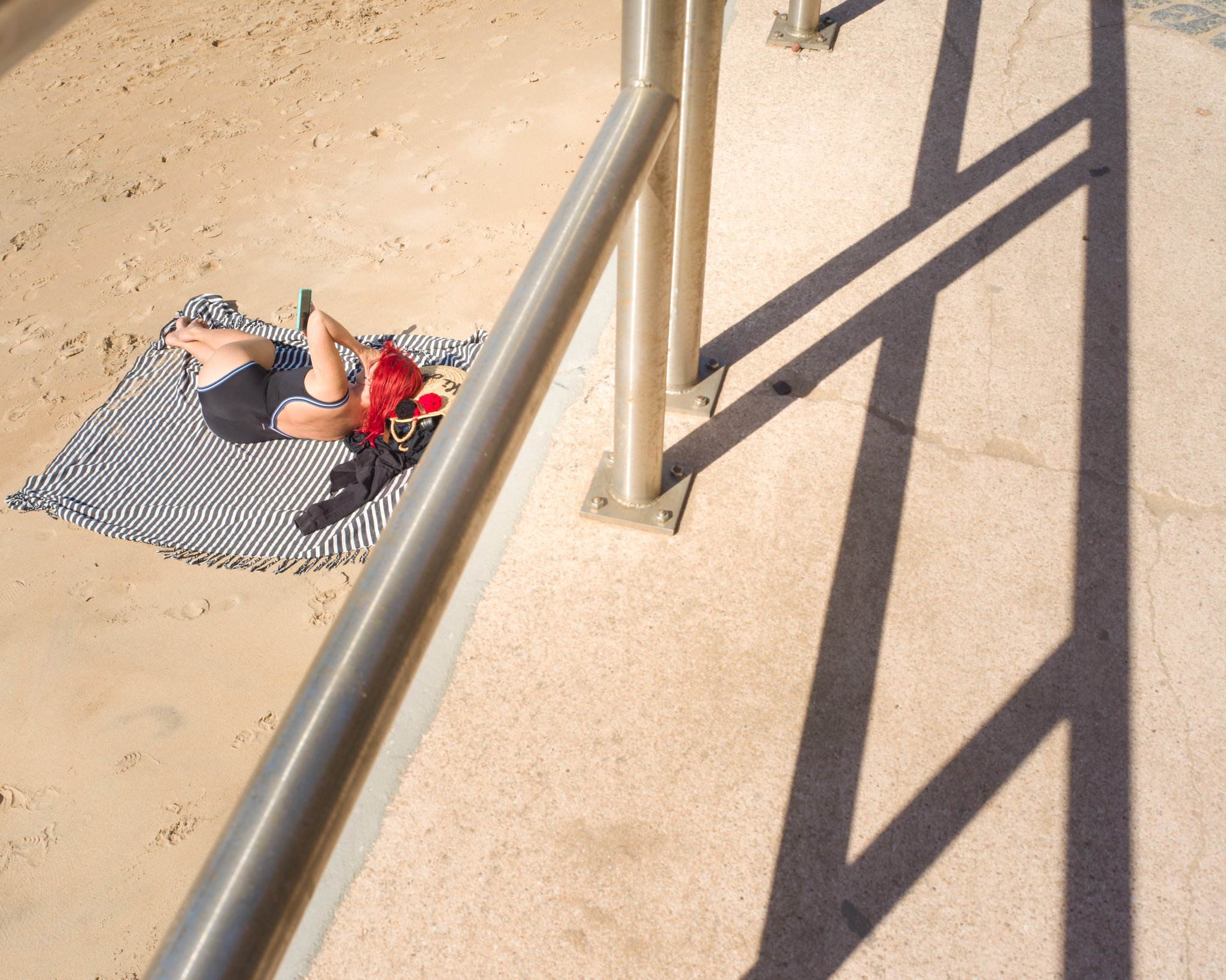 A woman with red hair sunbathing on a striped towel at the beach, wearing a black swimsuit, near a metal railing casting shadows on the sand.