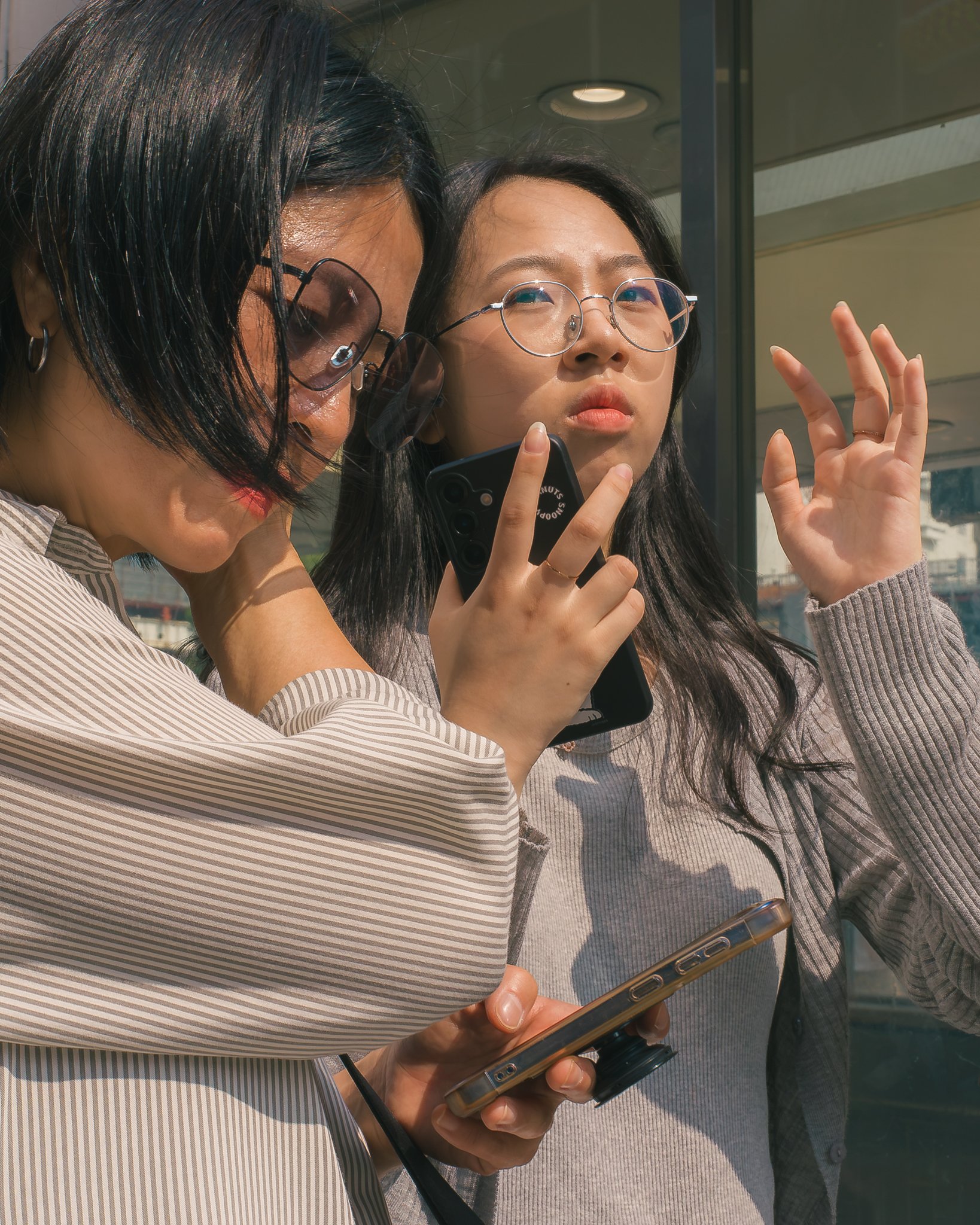 Two women, one with dark hair and glasses, the other with short dark hair and tinted glasses, are looking at and using a smartphone. They are standing next to a glass building, possibly at a transit station.